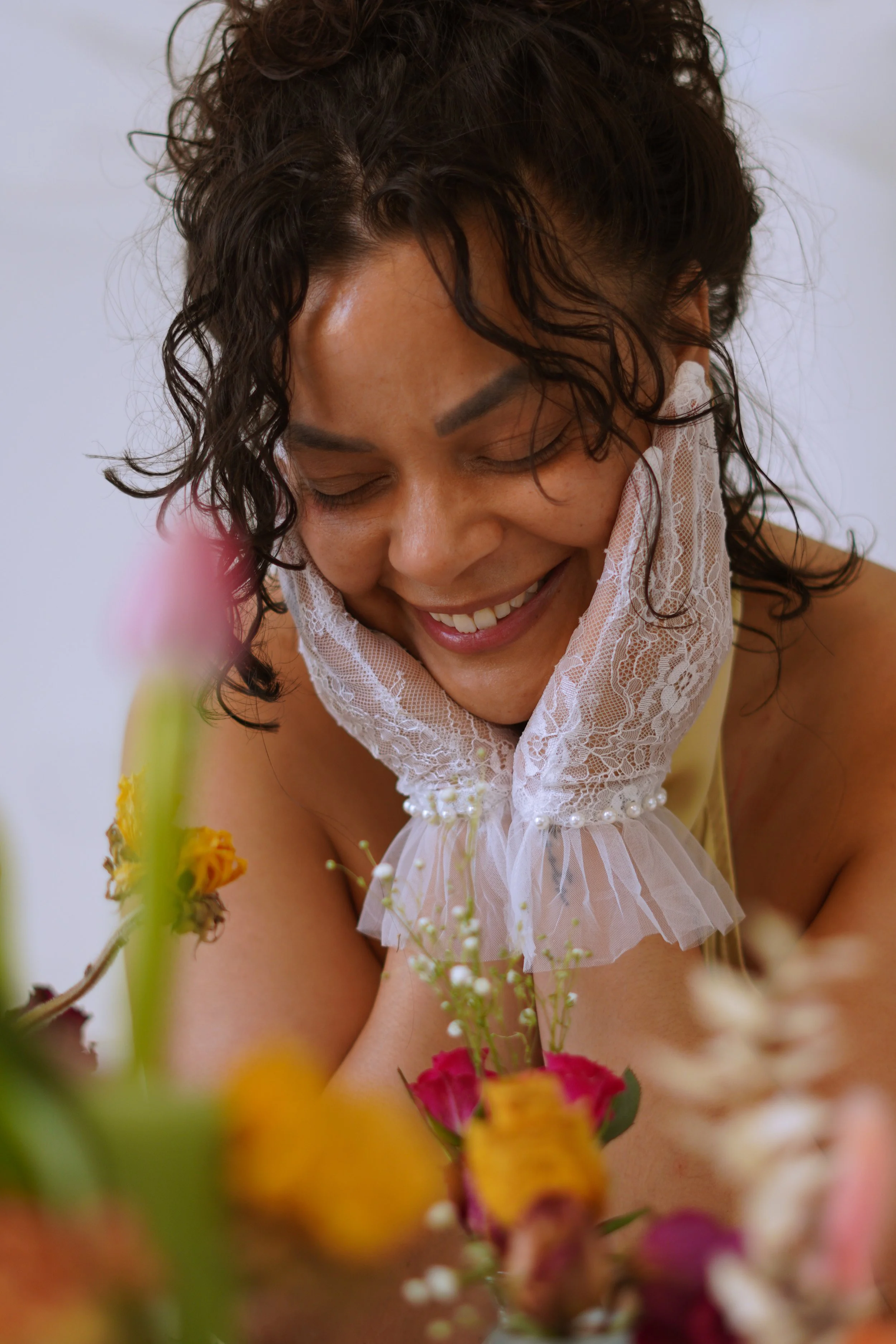 A woman with curly dark hair, smiling with her eyes closed, wearing a white lace neckpiece, surrounded by colorful flowers.