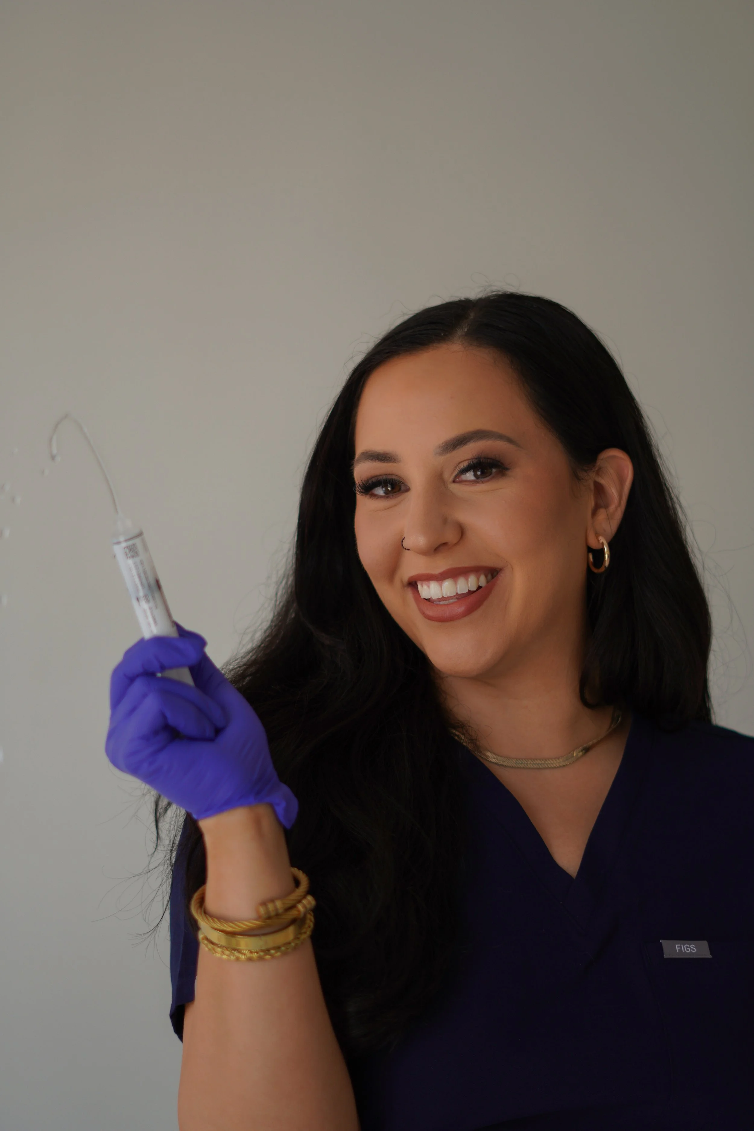 A smiling woman with dark hair, wearing gold jewelry and purple gloves, holding a syringe with a clear liquid against a plain gray background.