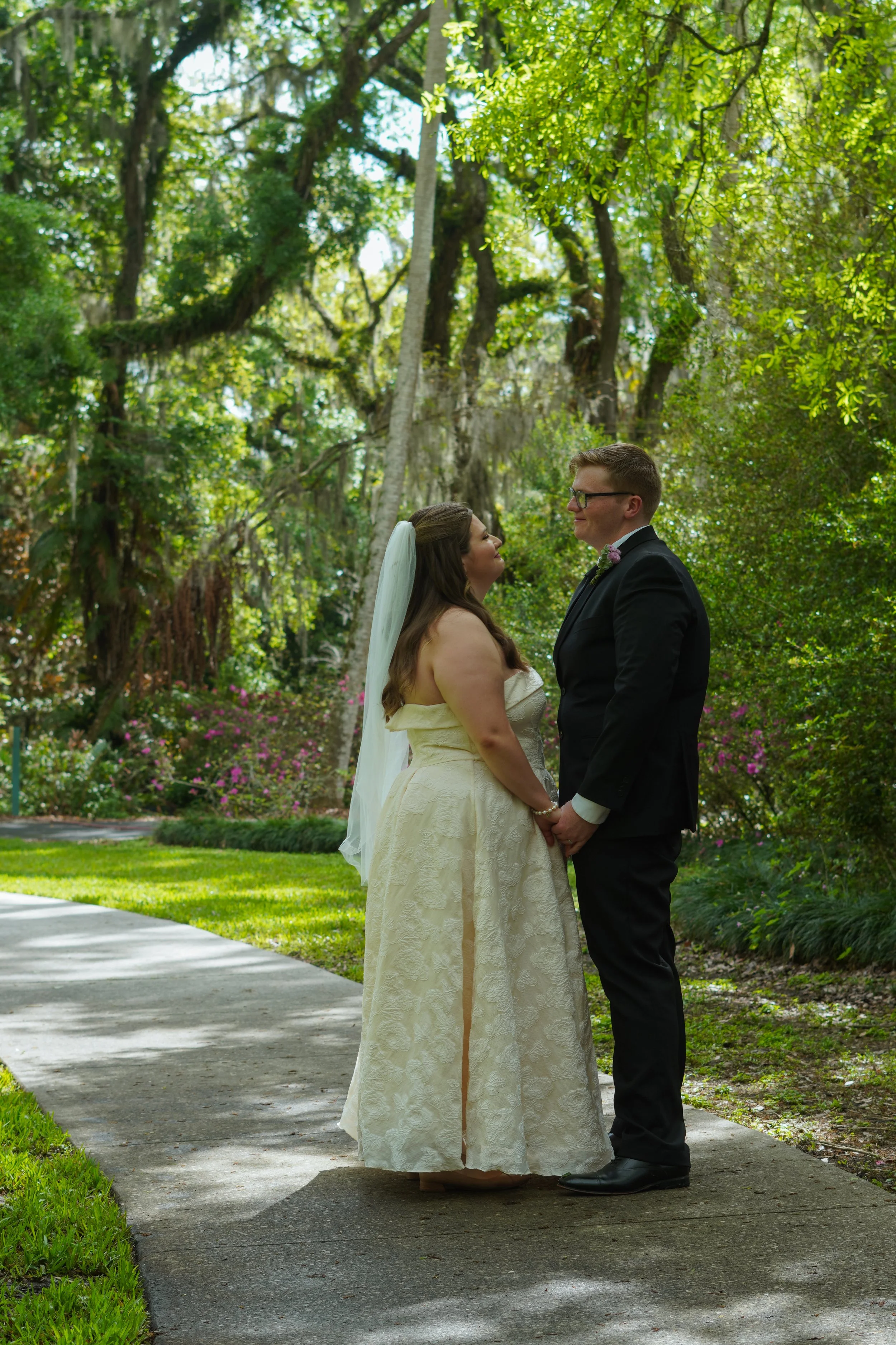 A bride and groom stand facing each other, holding hands, in a lush outdoor garden with tall trees and flowering bushes, during their wedding ceremony.