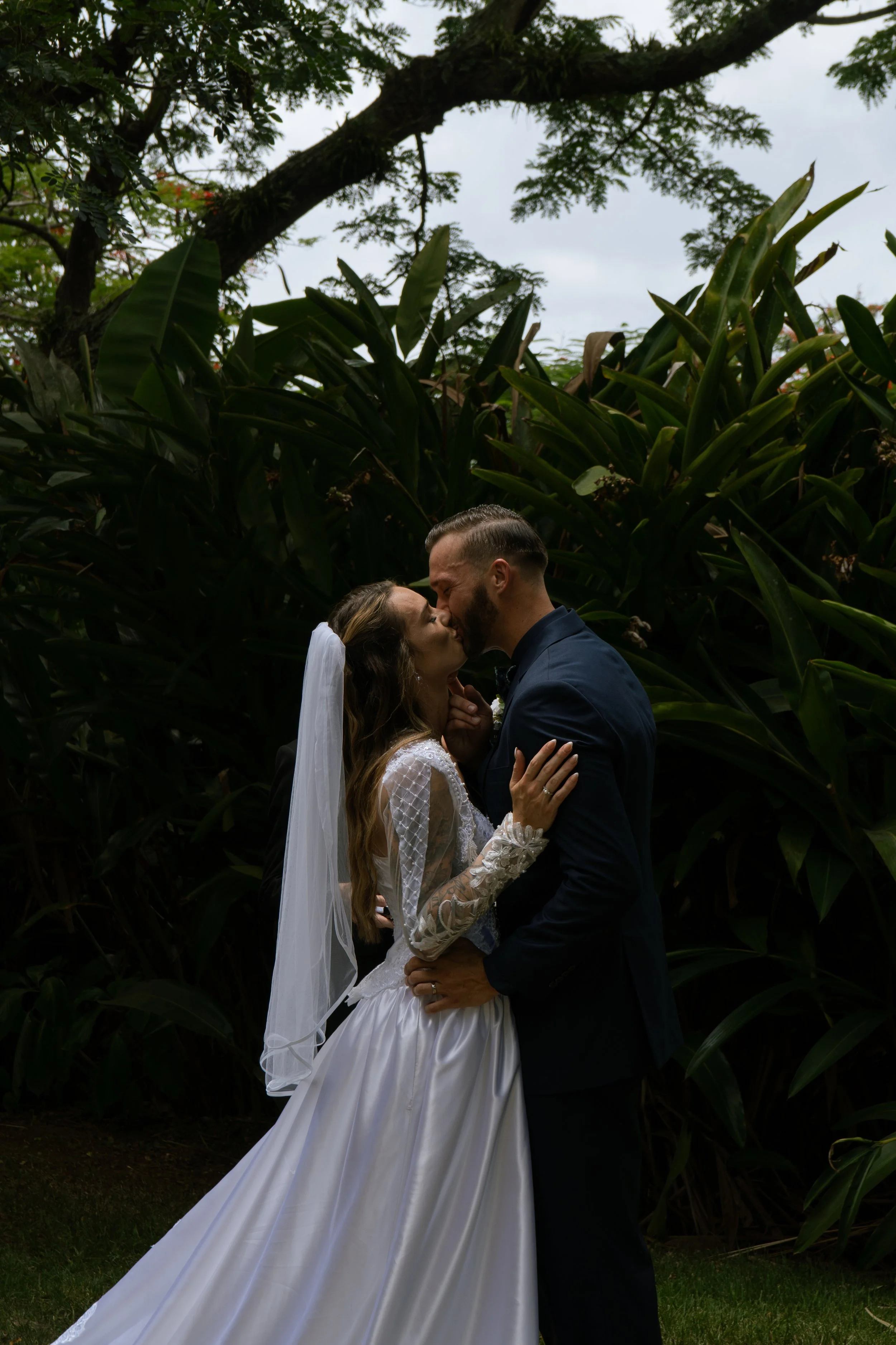 A newlywed couple sharing a kiss outdoors, surrounded by lush green trees and plants, on a cloudy day.