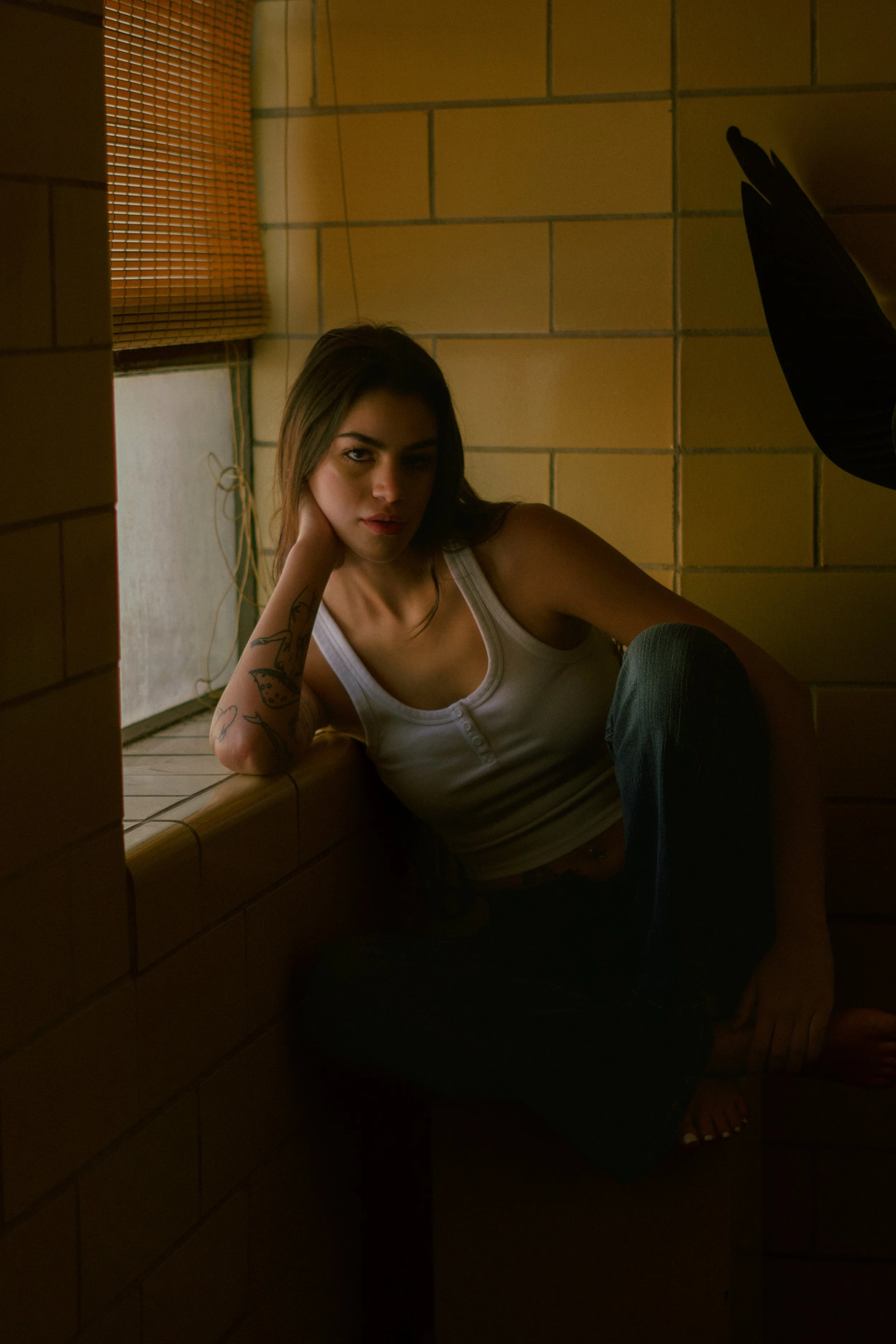A young woman with tattoos on her arm, sitting against a tiled wall near a window with a bamboo shade, looking at the camera with a relaxed expression.