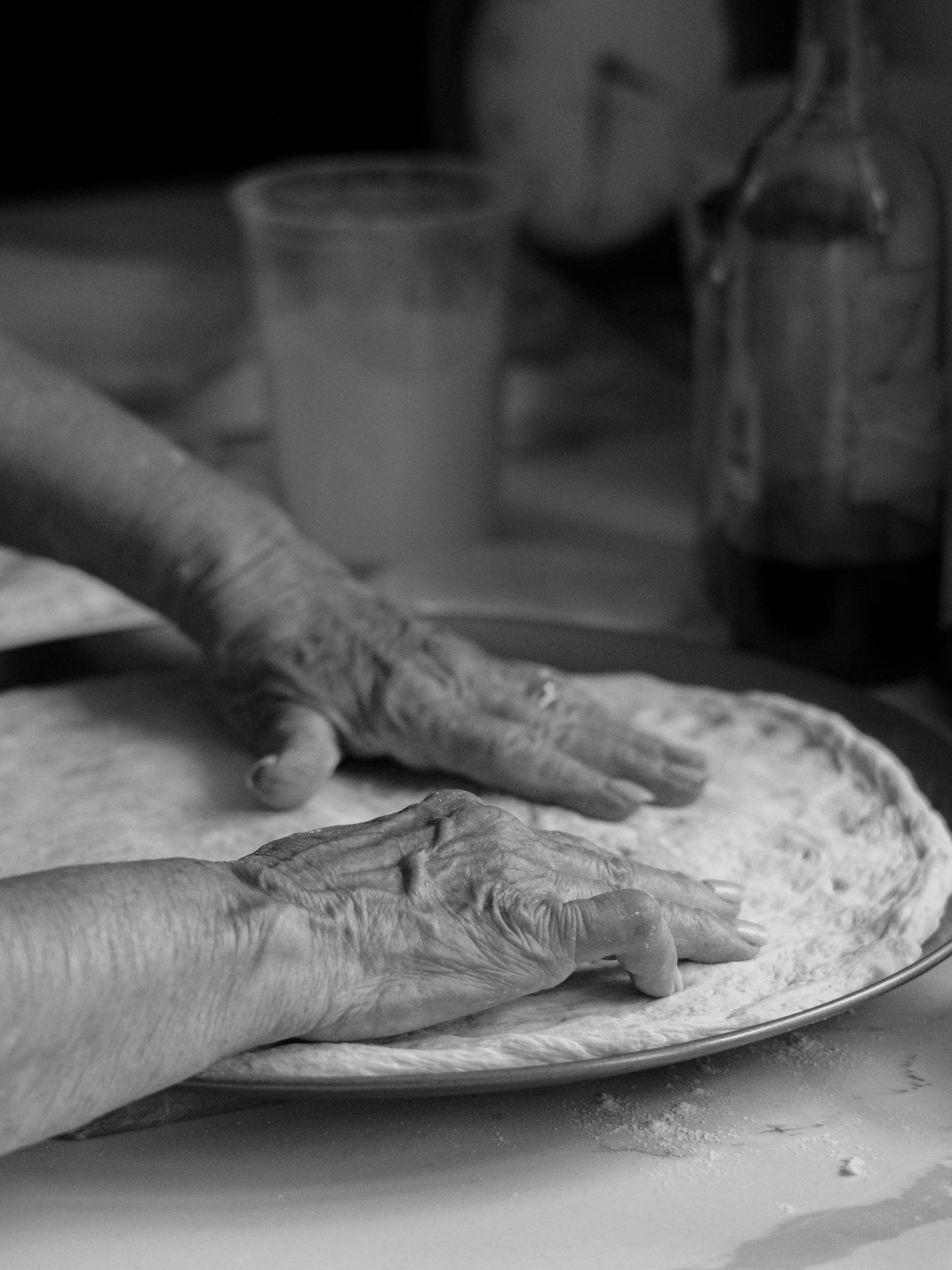 Close-up of a person's hand with textured skin, pressing dough on a baking tray, with other hands in the background preparing dough.