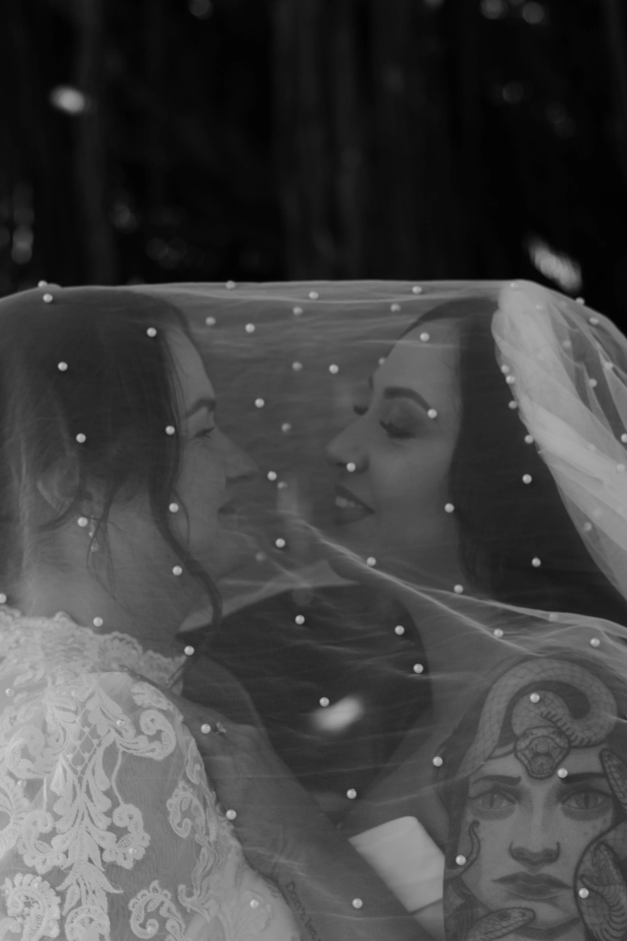 Two women dressed as brides kissing each other with their eyes closed, under a veil with pearl embellishments. One woman wears a lace wedding dress, and the other wears a sheer dress with floral embroidery.