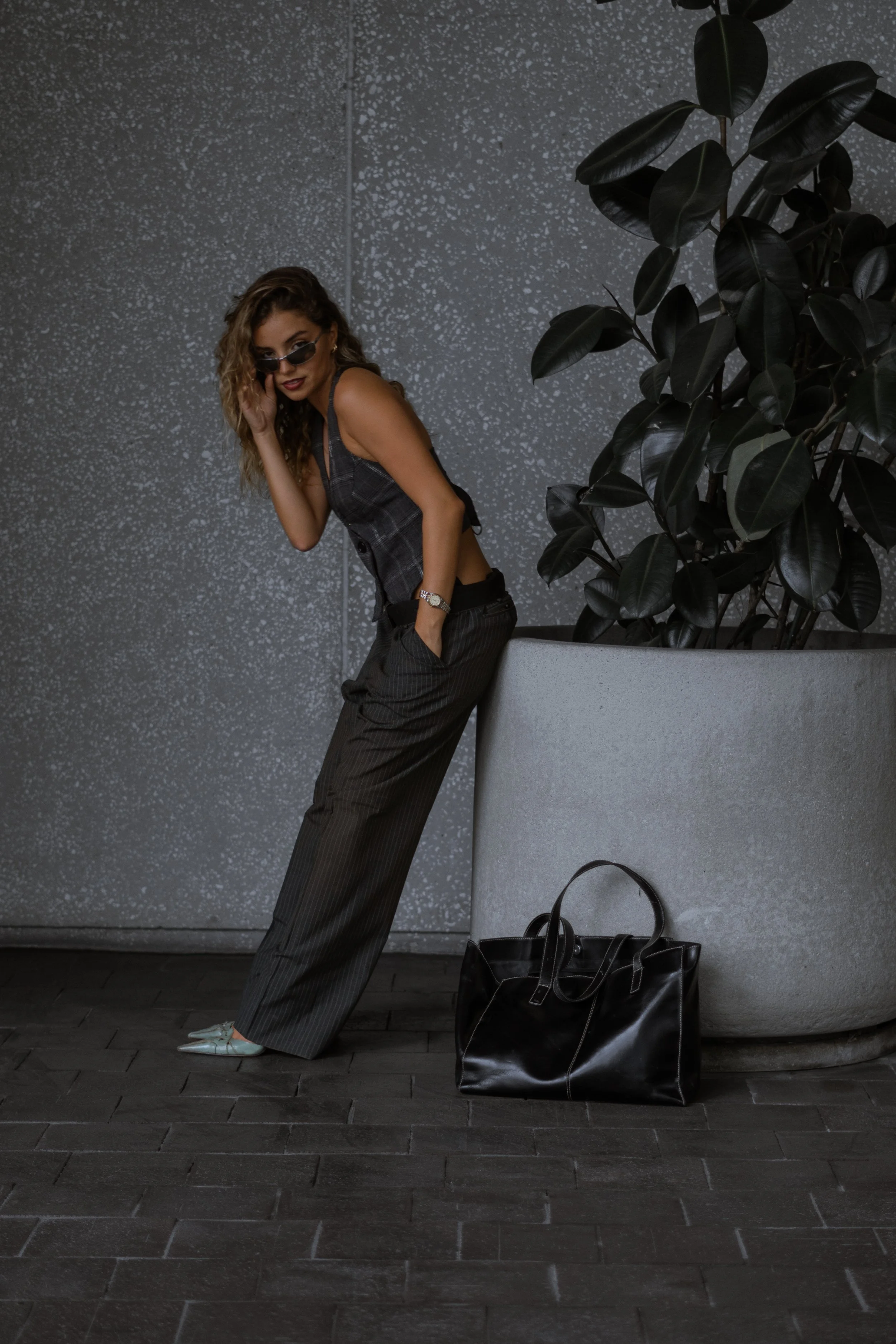 A woman with curly hair wearing sunglasses, a plaid sleeveless top, dark pinstripe pants, and white shoes, leaning against a textured gray wall next to a large black handbag on the ground and a large potted plant with dark green leaves.