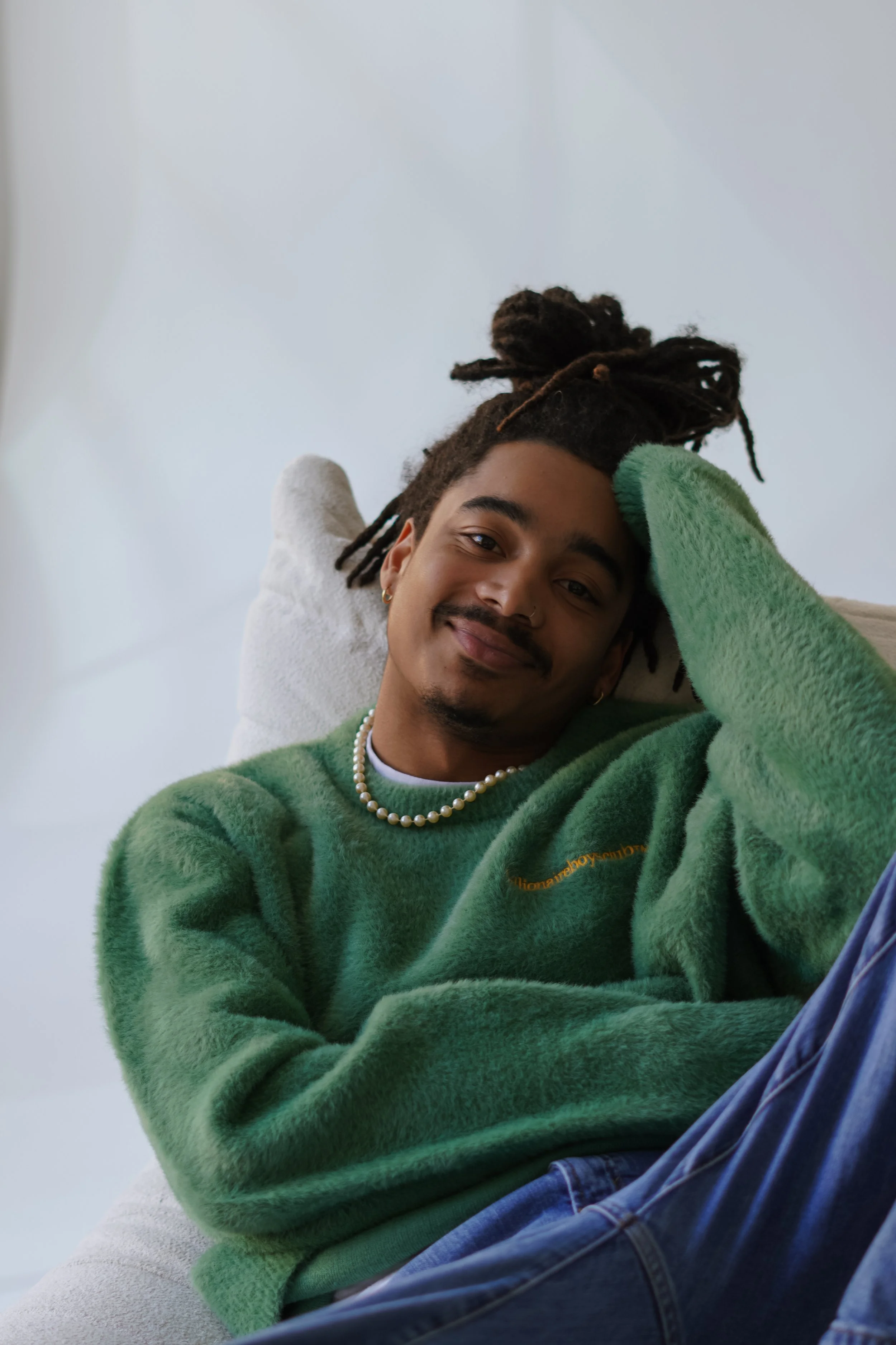 A young man with dark dreadlocks, smiling, wearing a green fuzzy sweater, a pearl necklace, and gold earrings, sitting against a plain light-colored wall.