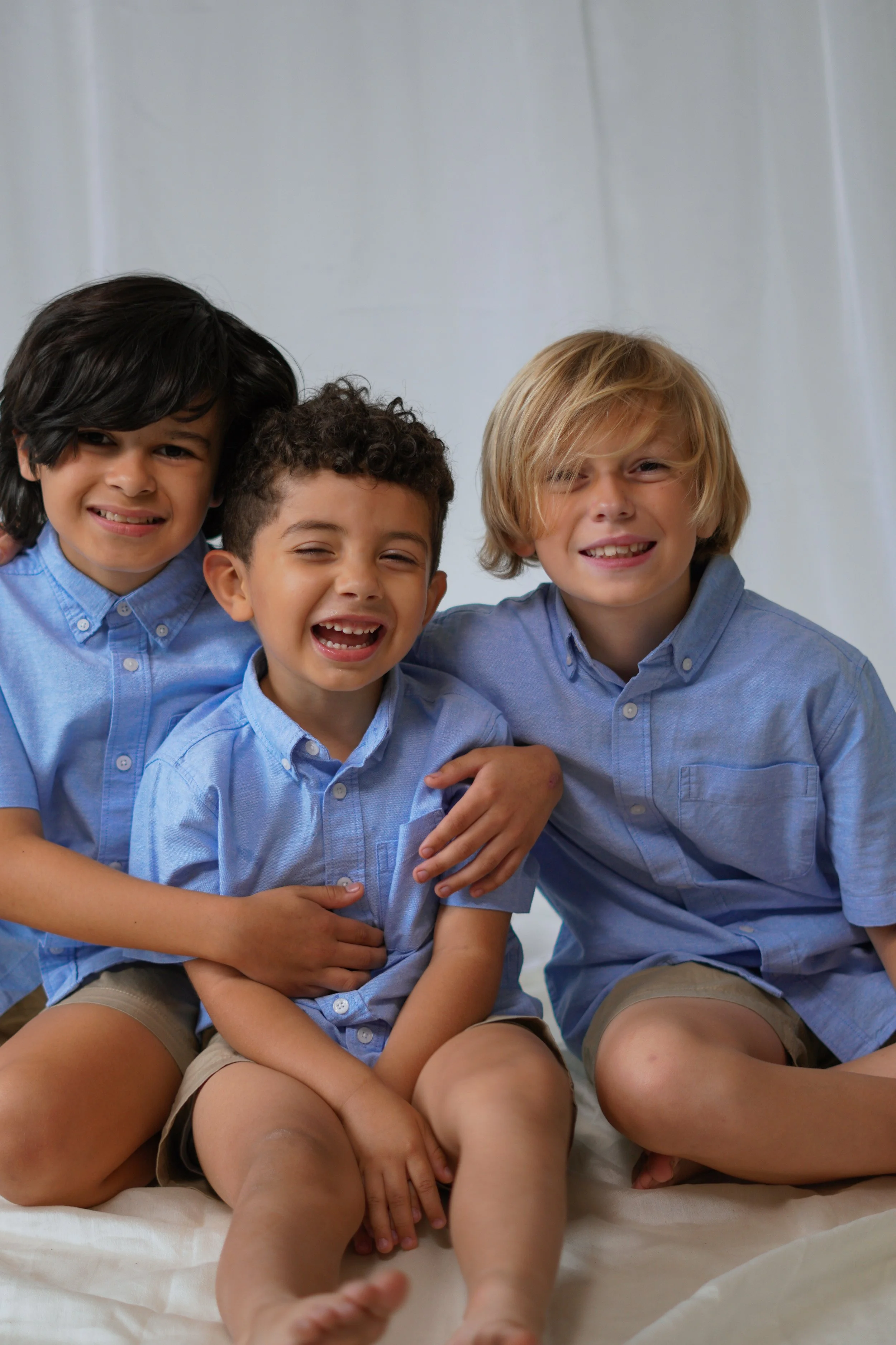 Three boys sitting together, smiling and laughing, wearing light blue button-up shirts and khaki shorts, against a plain background.