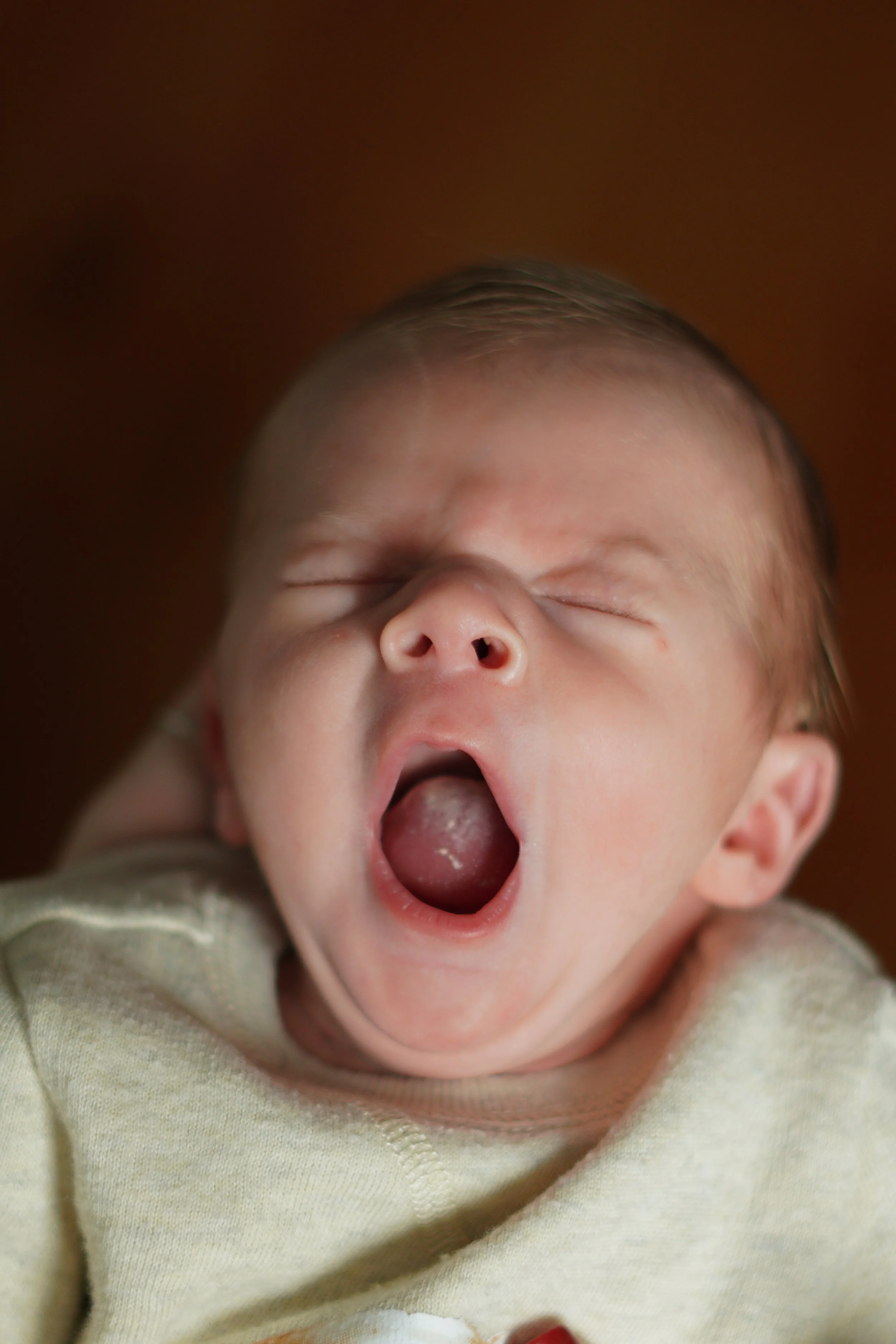 Close-up of a baby with eyes closed, mouth open, yawning, wearing a light yellow shirt.