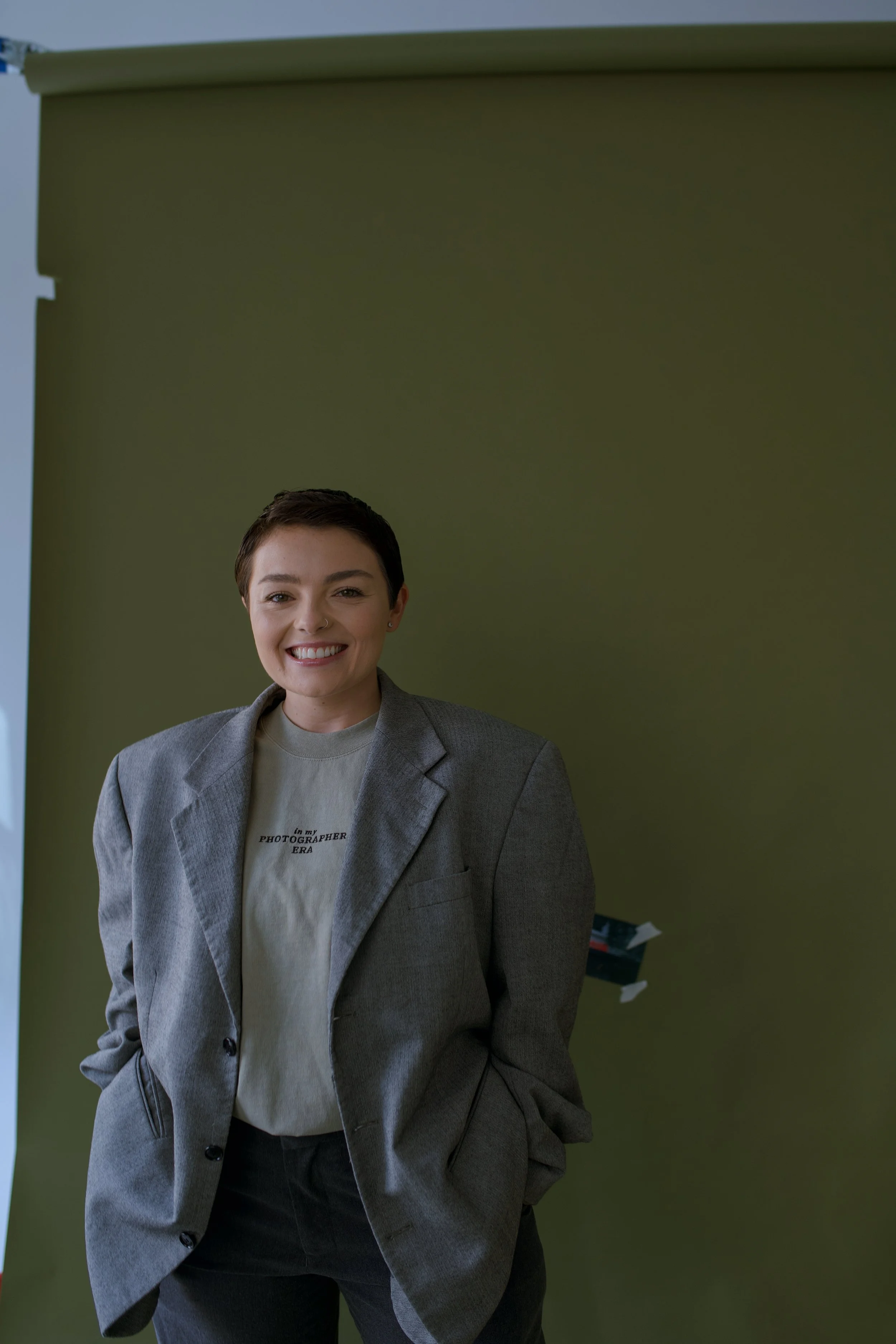 A woman with short dark hair smiling, dressed in a gray blazer and casual T-shirt, standing in front of a green backdrop.