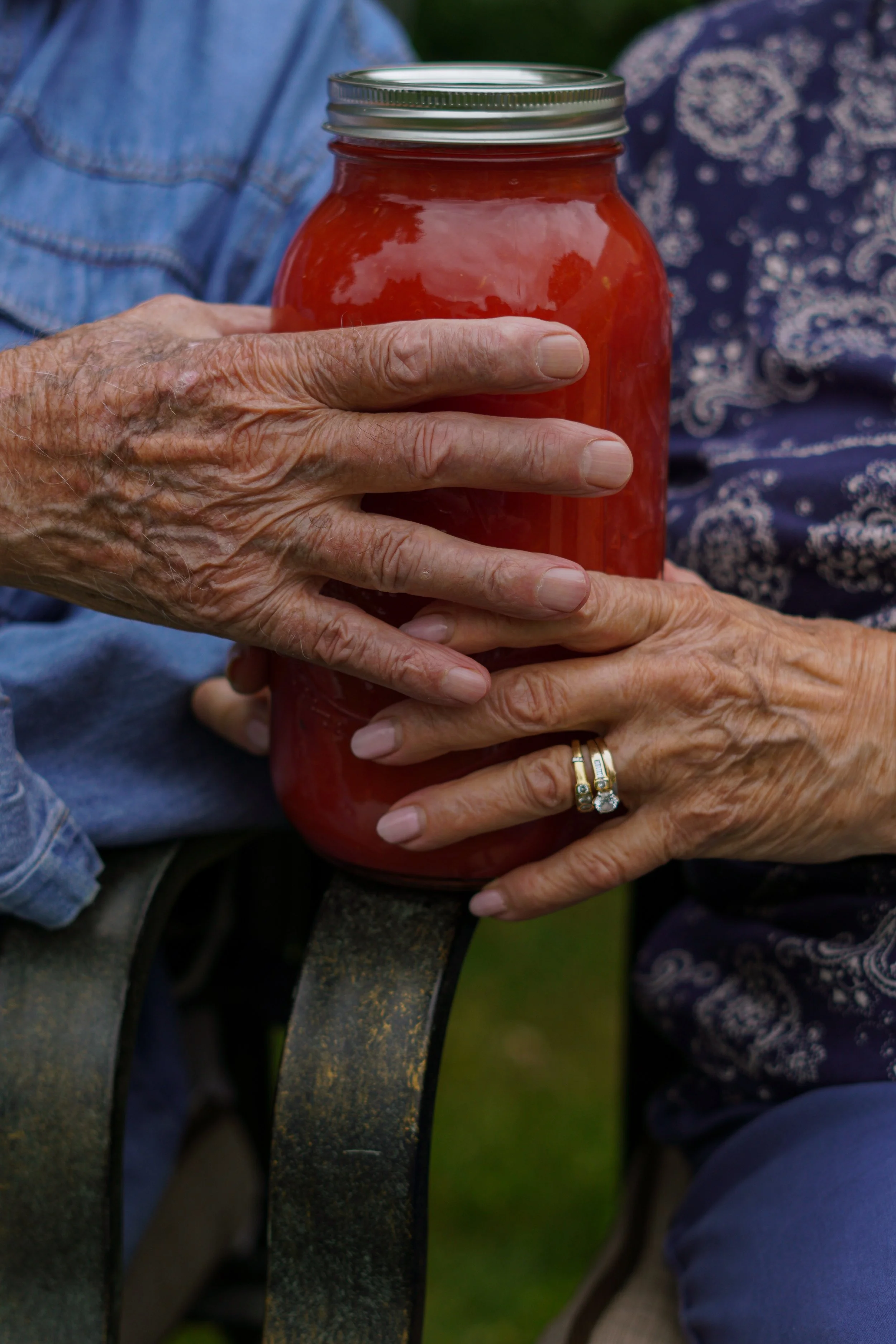 Close-up of elderly hands holding a glass jar filled with red preserves or jam, with some visible fruit pieces inside. The person wears a gold ring with a large gemstone and a wedding band on the ring finger.