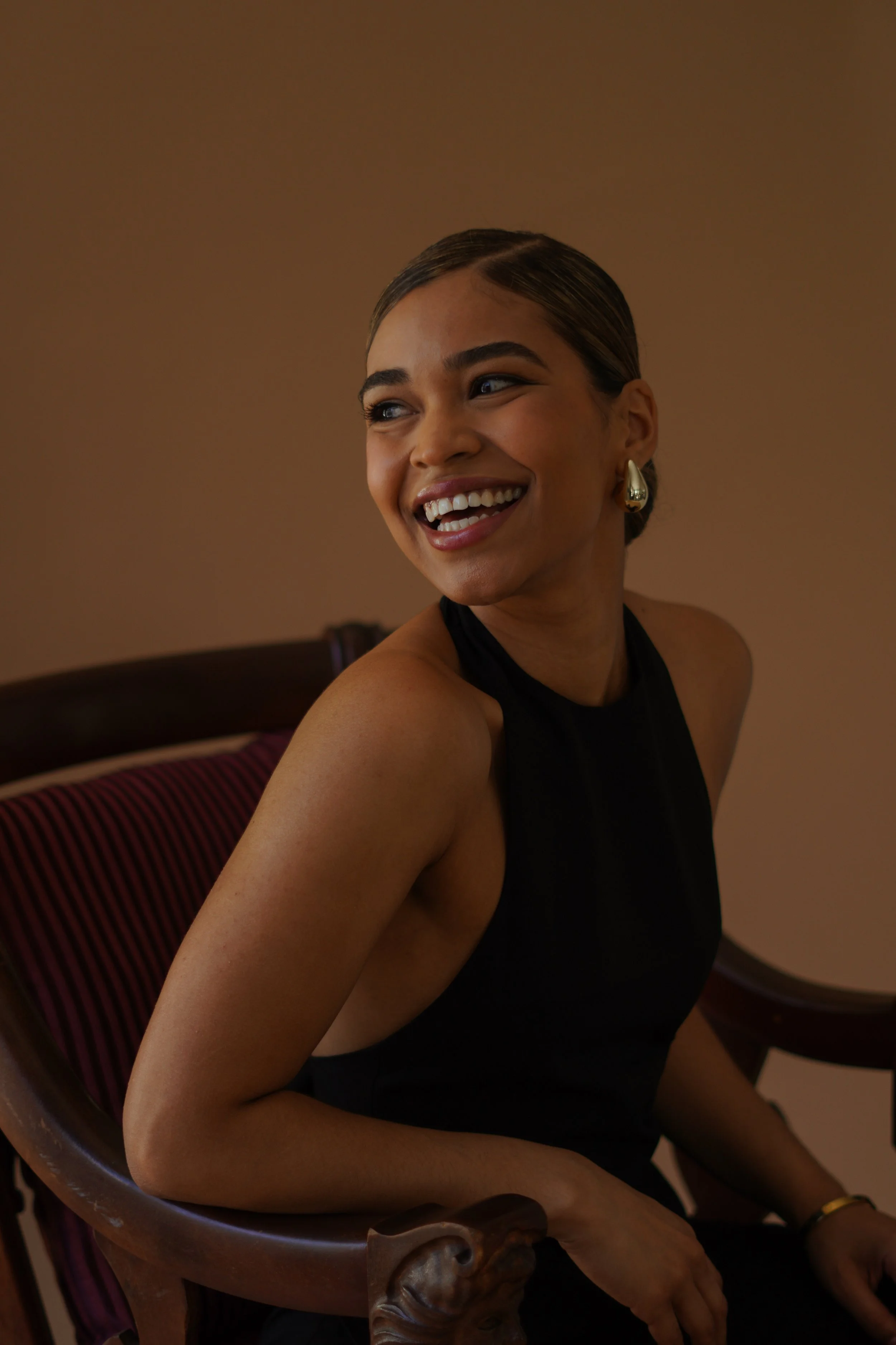 A woman with dark hair in a low bun, wearing large gold earrings and a sleeveless black top, smiling while sitting in a wooden chair with a striped cushion, against a plain brown background.