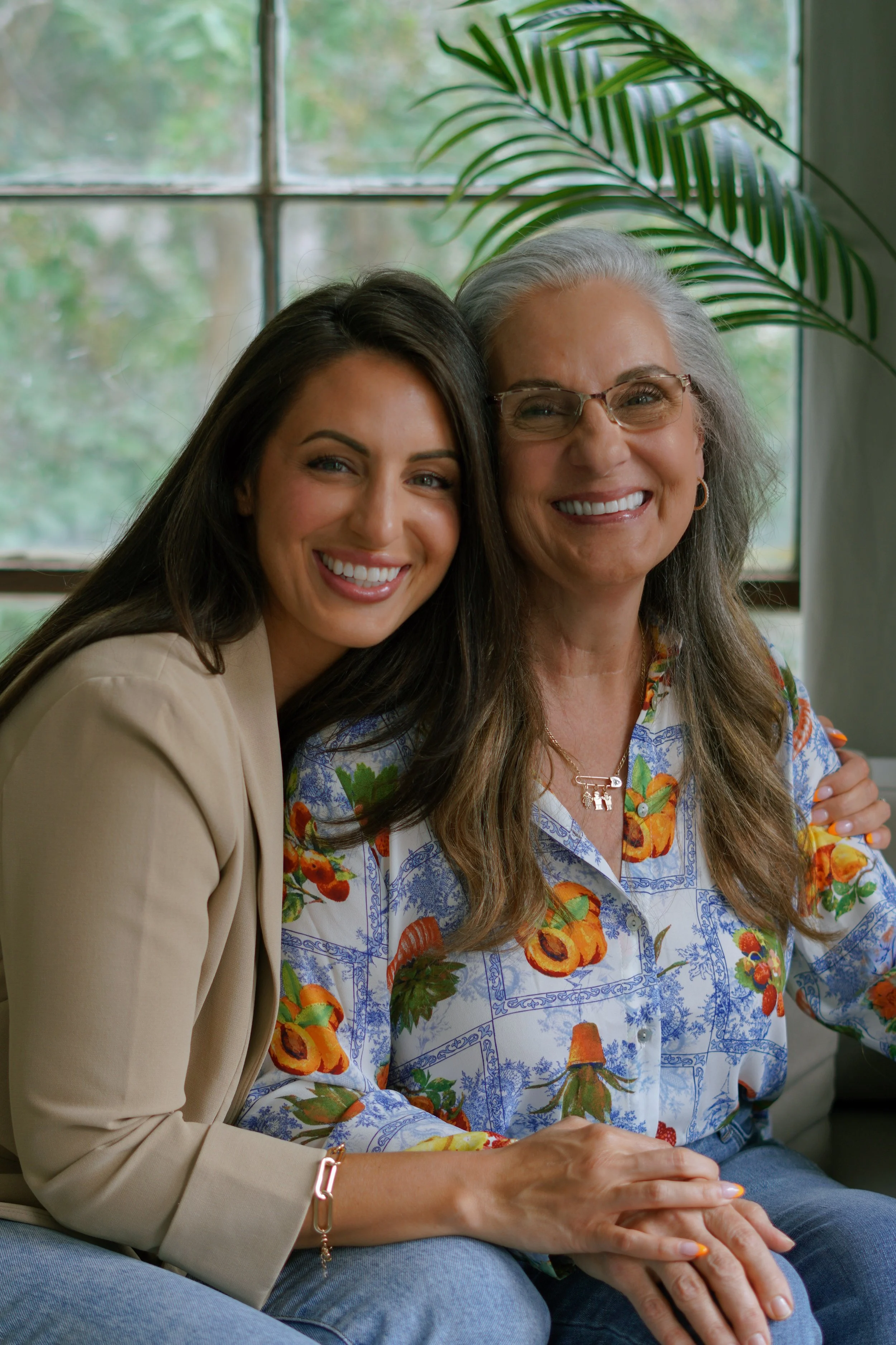 Two women sitting closely together in front of a window, smiling at the camera. One is an older woman with gray hair and glasses, wearing a colorful floral blouse. The other is a younger woman with long brown hair, wearing a beige blazer. There is a 