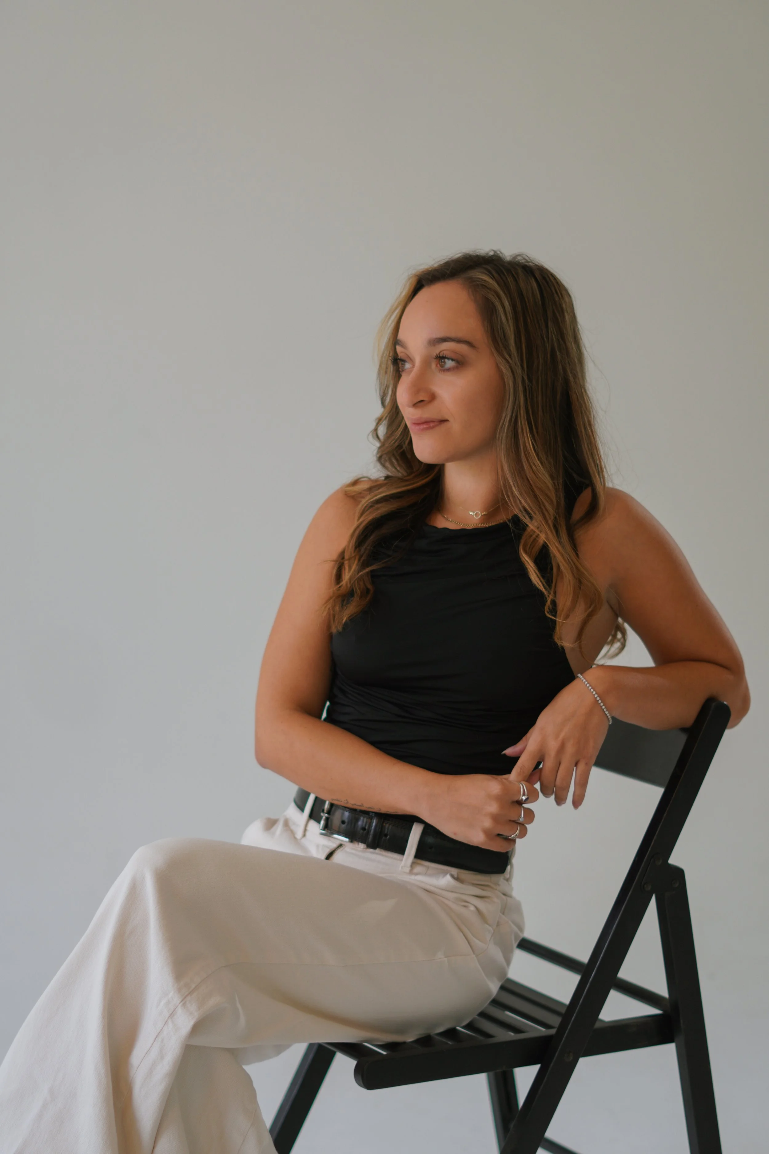 A woman with long wavy brown hair sitting on a black folding chair, wearing a black sleeveless top and beige pants, looking to her right, with a neutral expression, against a plain light gray background.