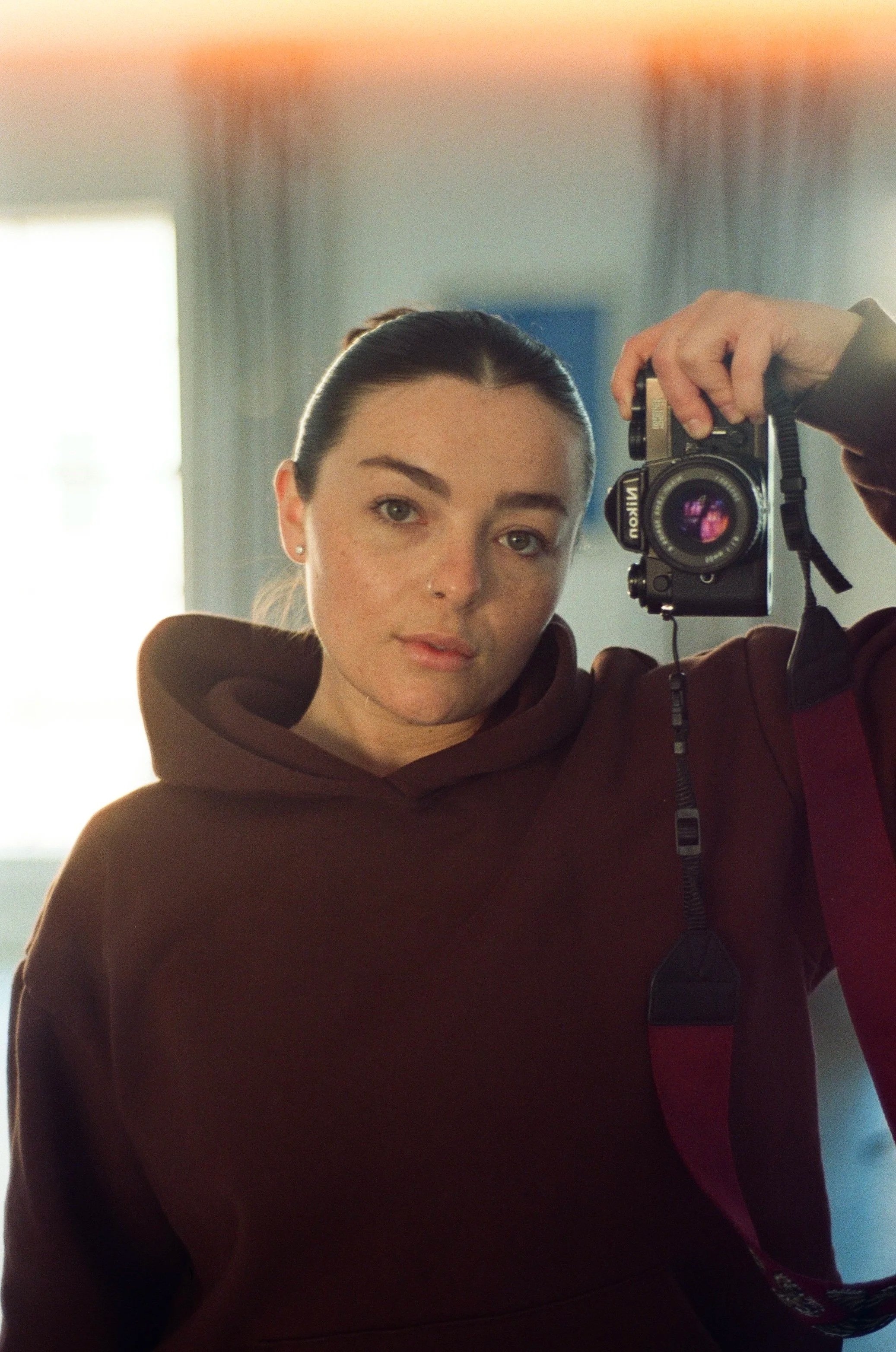 A woman with dark hair tied back, wearing earrings and a brown hoodie, is taking a selfie in front of a mirror with a camera. The background shows a bright window with curtains, and a softly lit room.