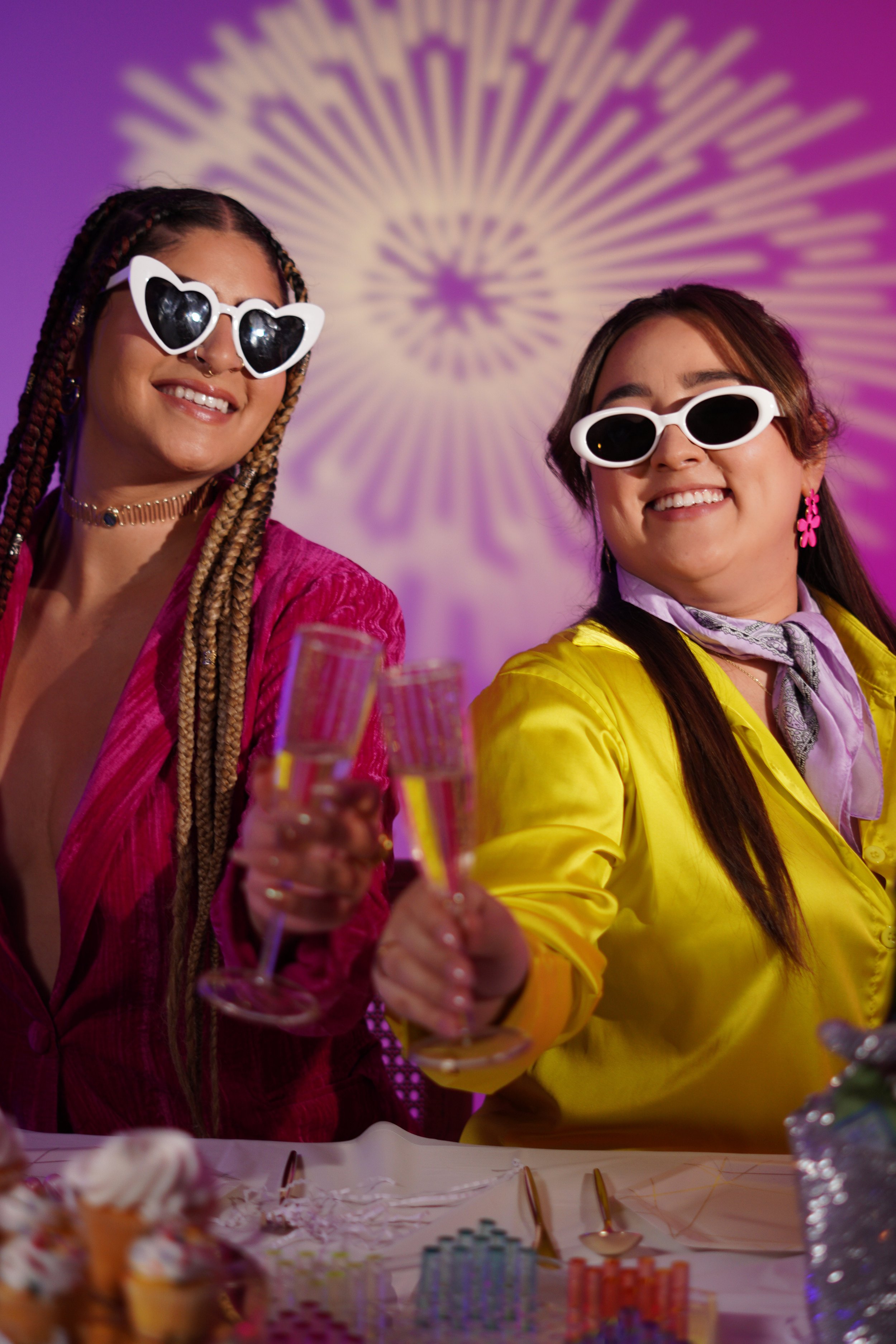 Two women at a celebration, wearing colorful outfits and white heart-shaped sunglasses, smiling and clinking champagne glasses. The background features colorful lighting and decorations.