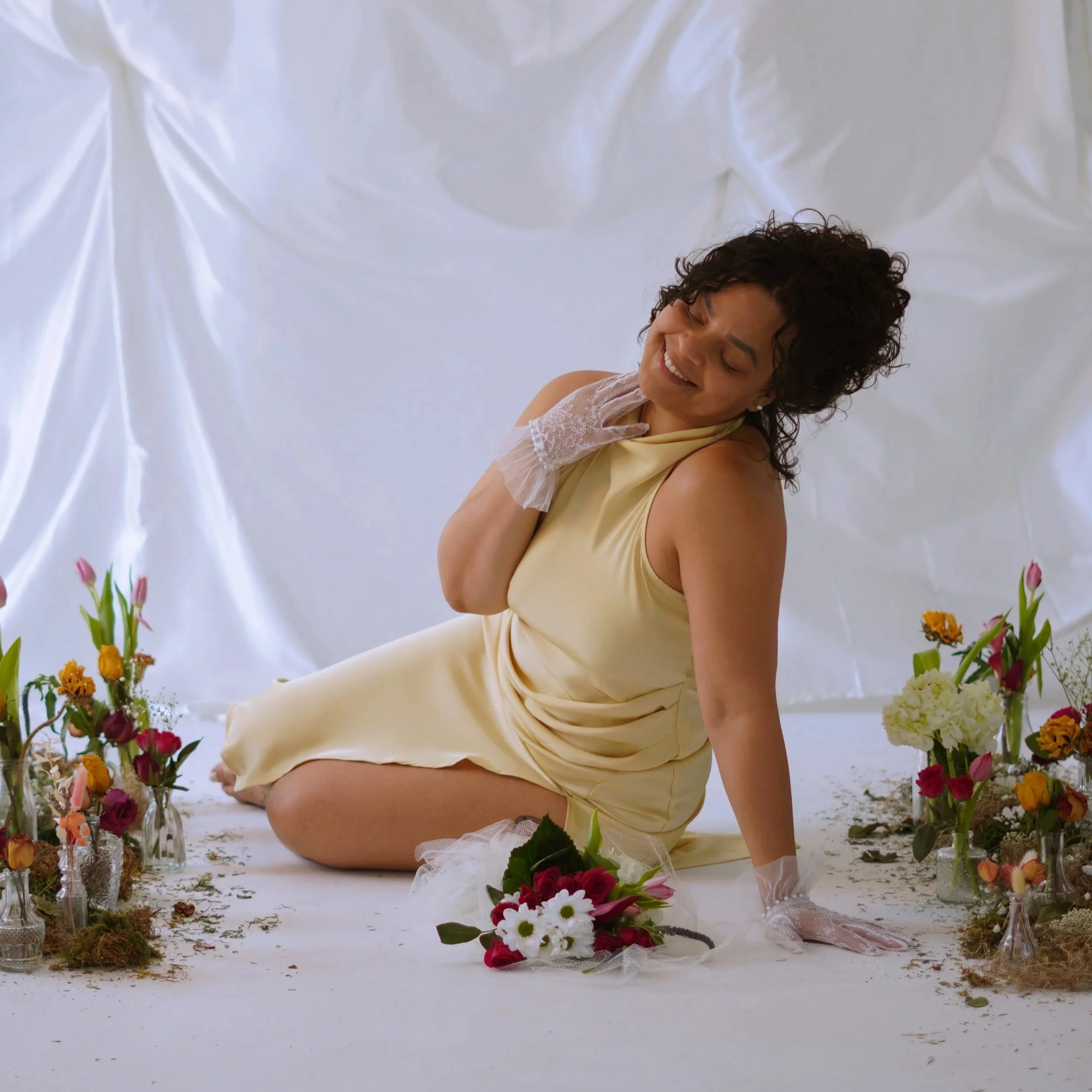 A woman smiling and lying on the floor among colorful flowers and glass vases, set against a white fabric backdrop.
