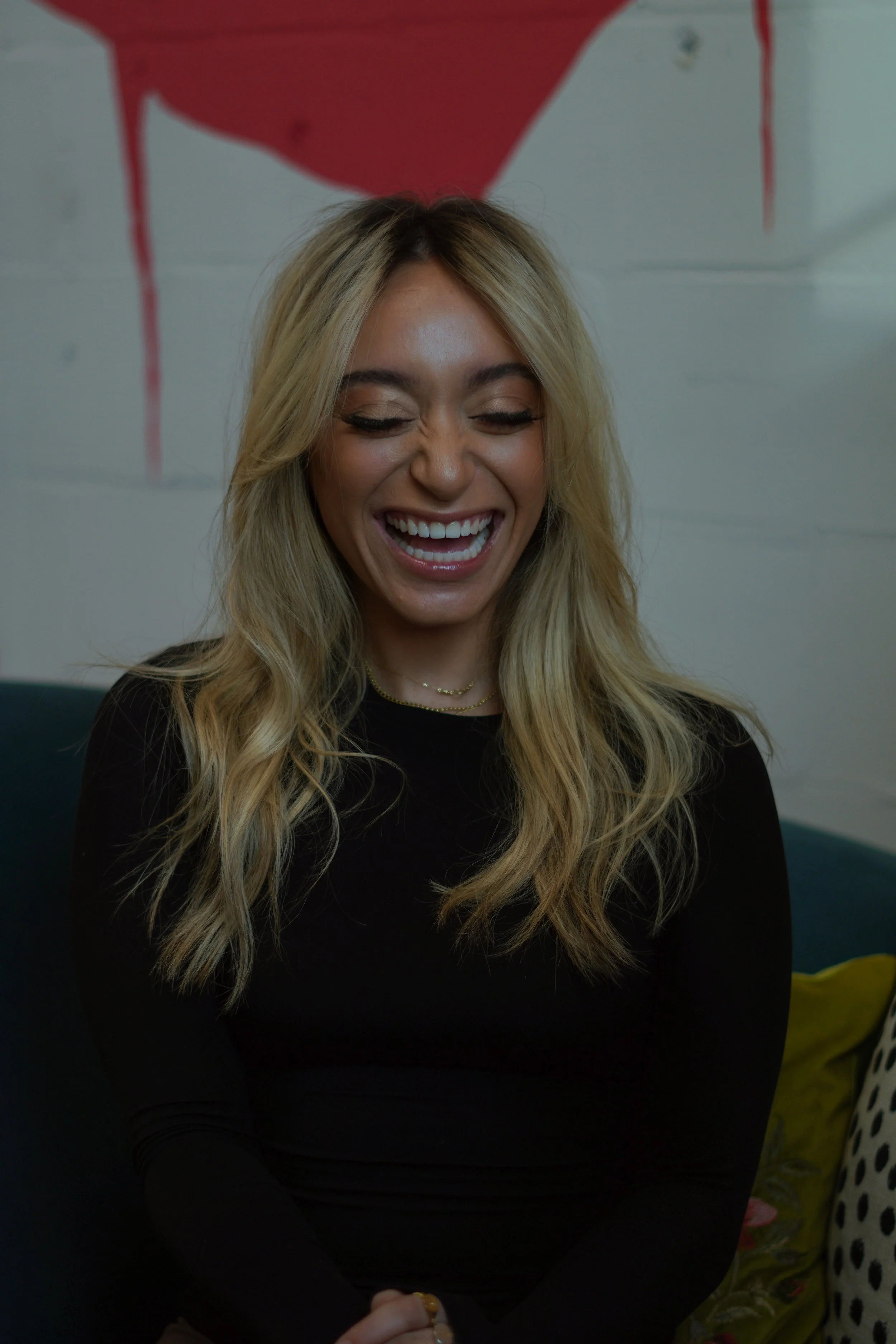 A woman with long blonde hair smiling and showing teeth, wearing a black top and layered necklaces, sitting against a painted white brick wall with red paint drips.
