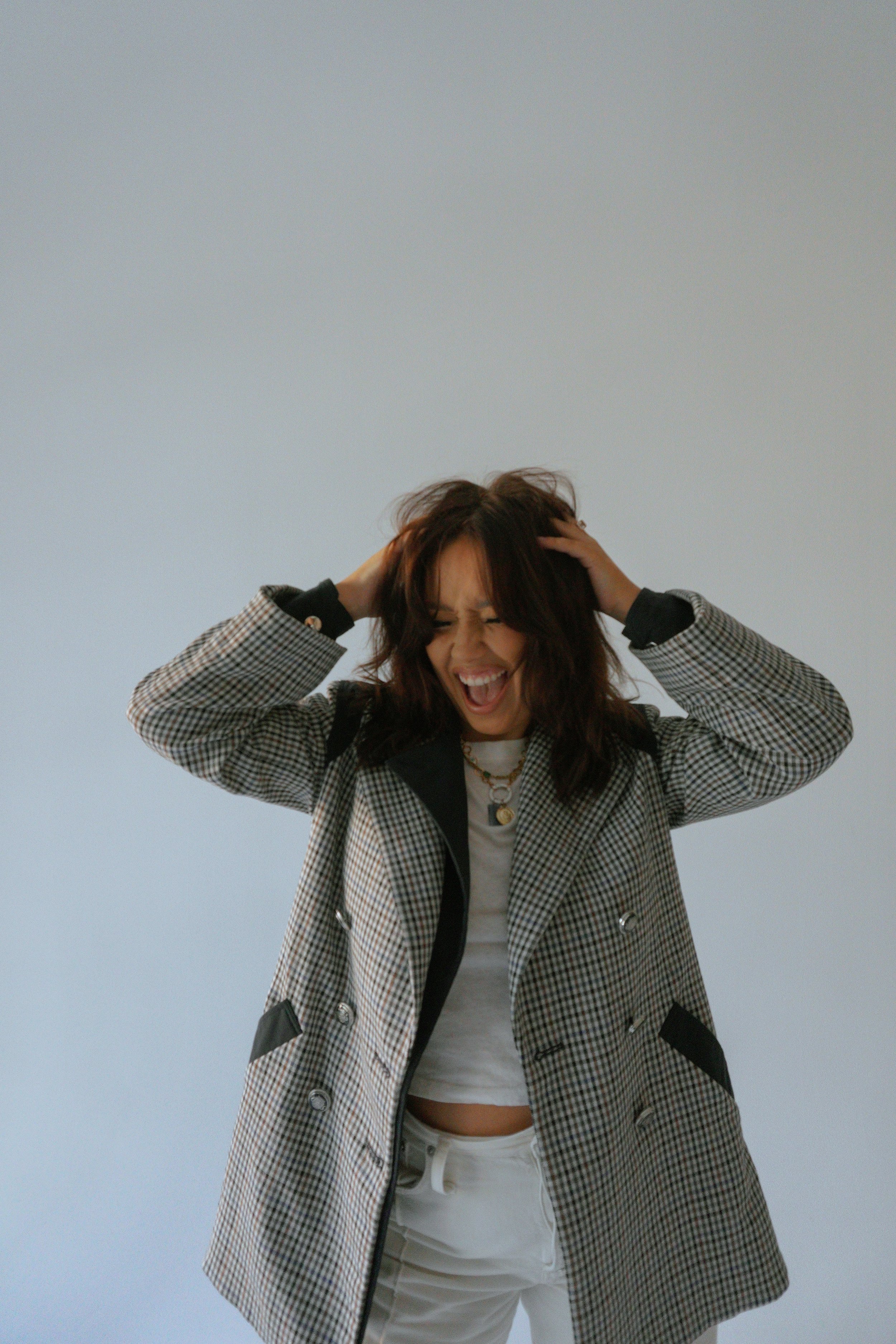 A woman with brown hair is smiling and holding her head with both hands. She is wearing a checkered blazer, a white crop top, white pants, and layered necklaces.