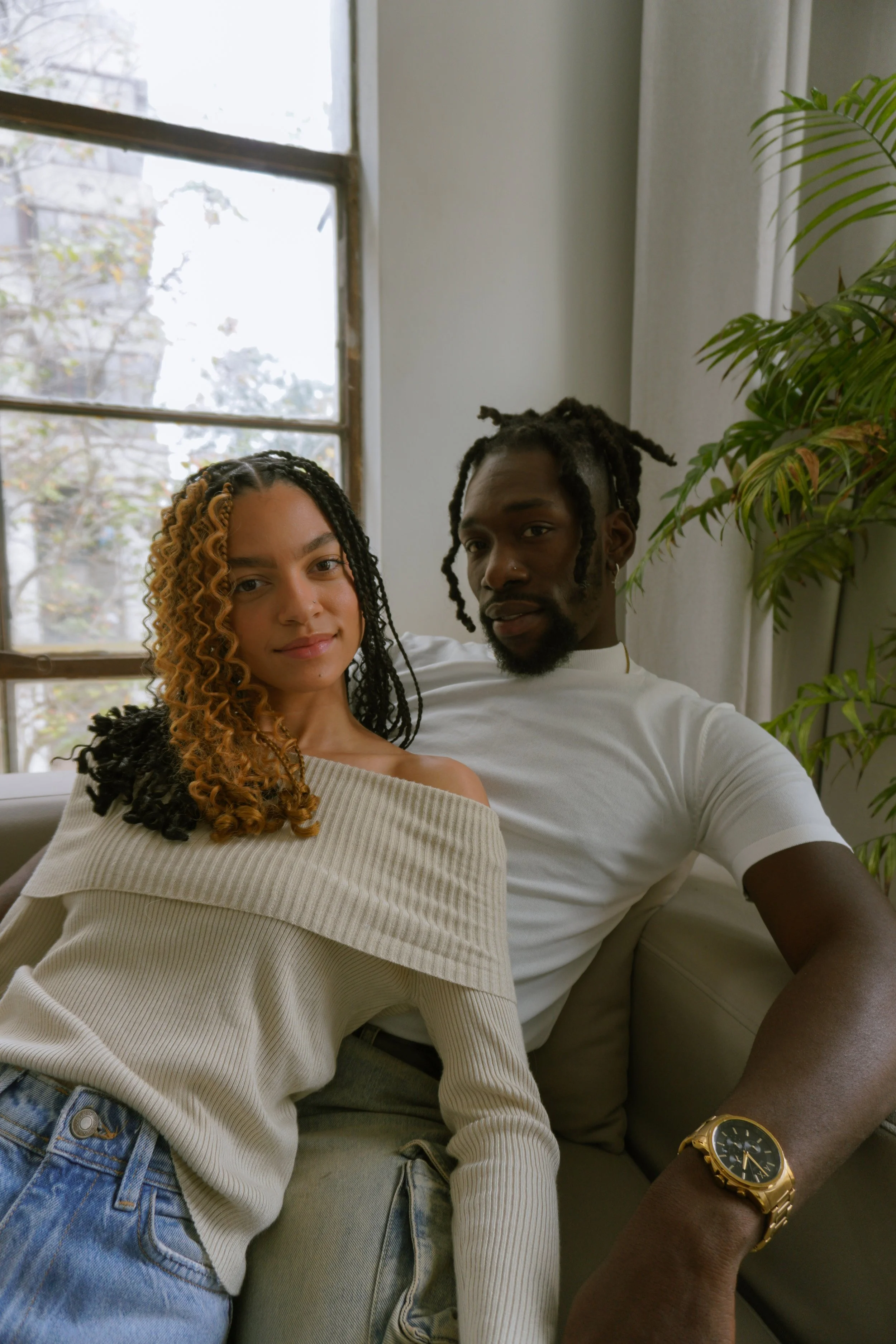 A young woman and a young man sitting together indoors in front of a window and a houseplant. The woman has partly curly hair with blonde tips and is wearing a beige off-shoulder sweater and jeans. The man has dreadlocks, a nose ring, and is wearing 