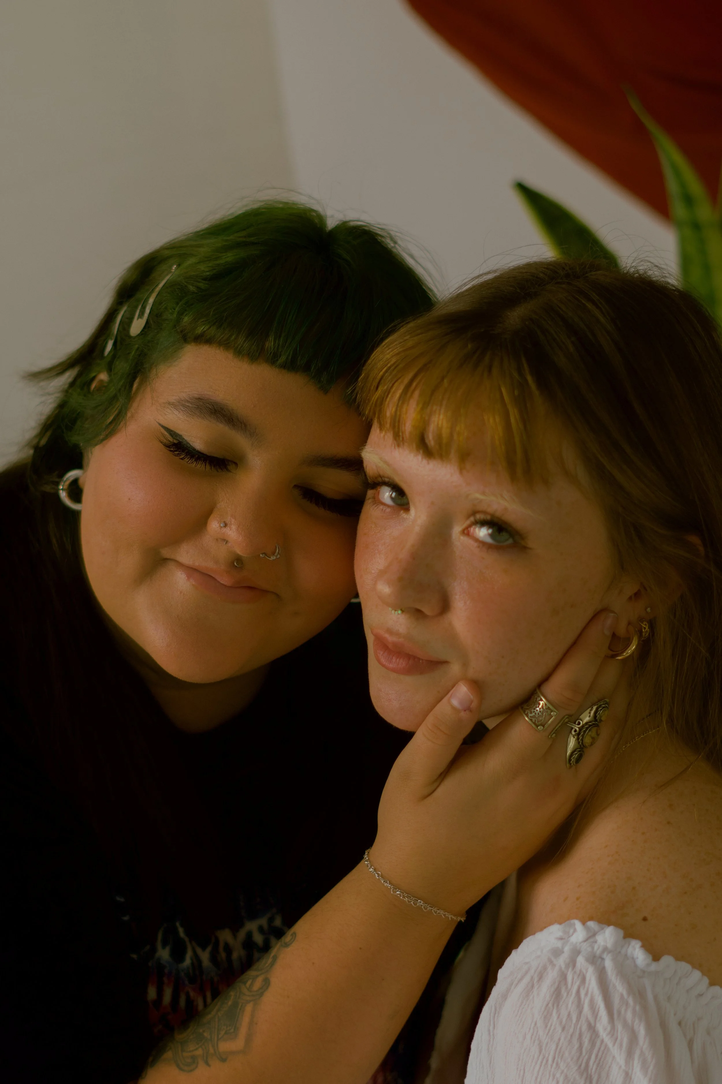 Two women, one with green hair and the other with red hair, close together, smiling, with the red-haired woman touching her face and showing jewelry including rings, earrings, and a bracelet.