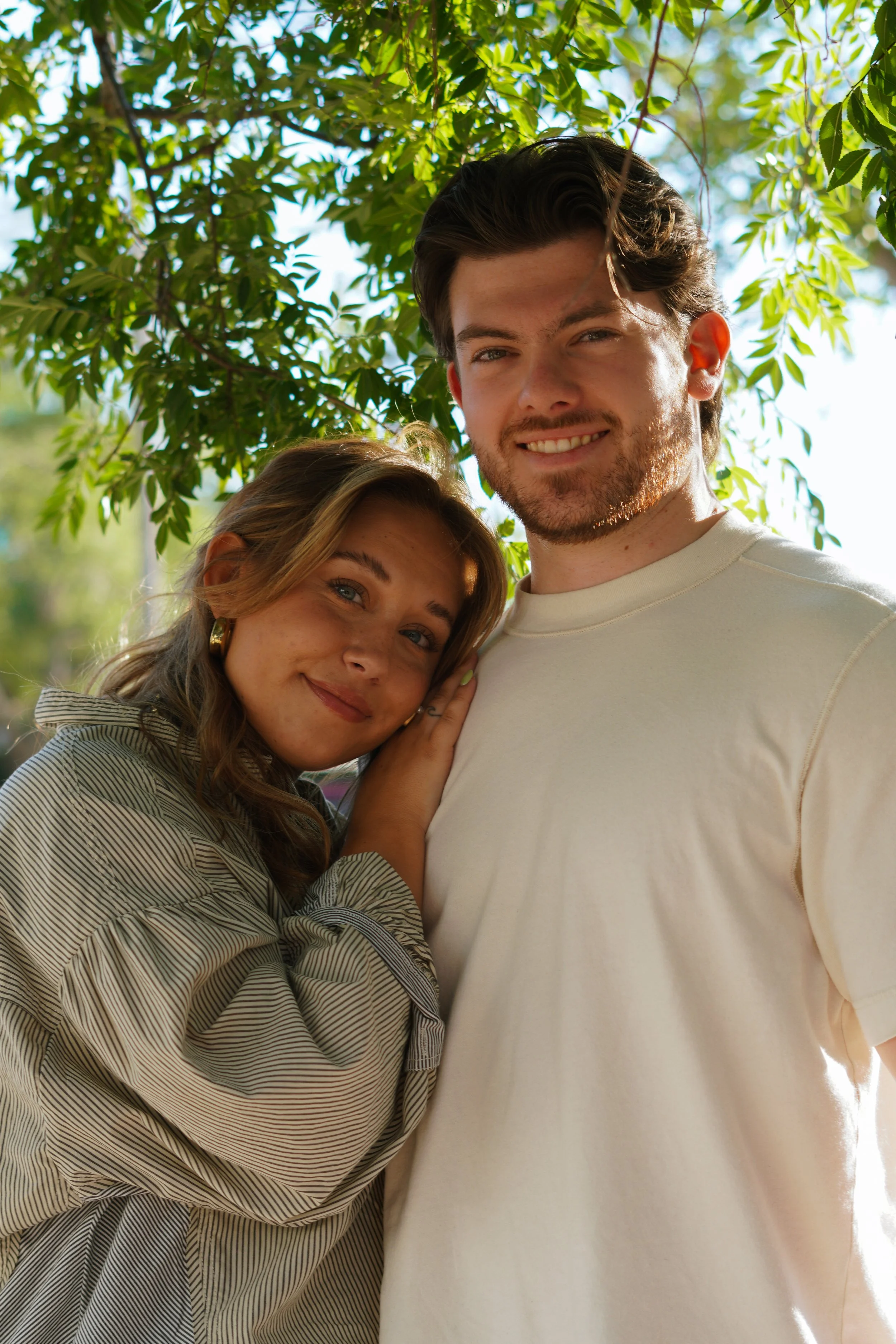 A young woman with blonde hair and a young man with dark hair standing outdoors under a leafy tree, smiling at the camera.