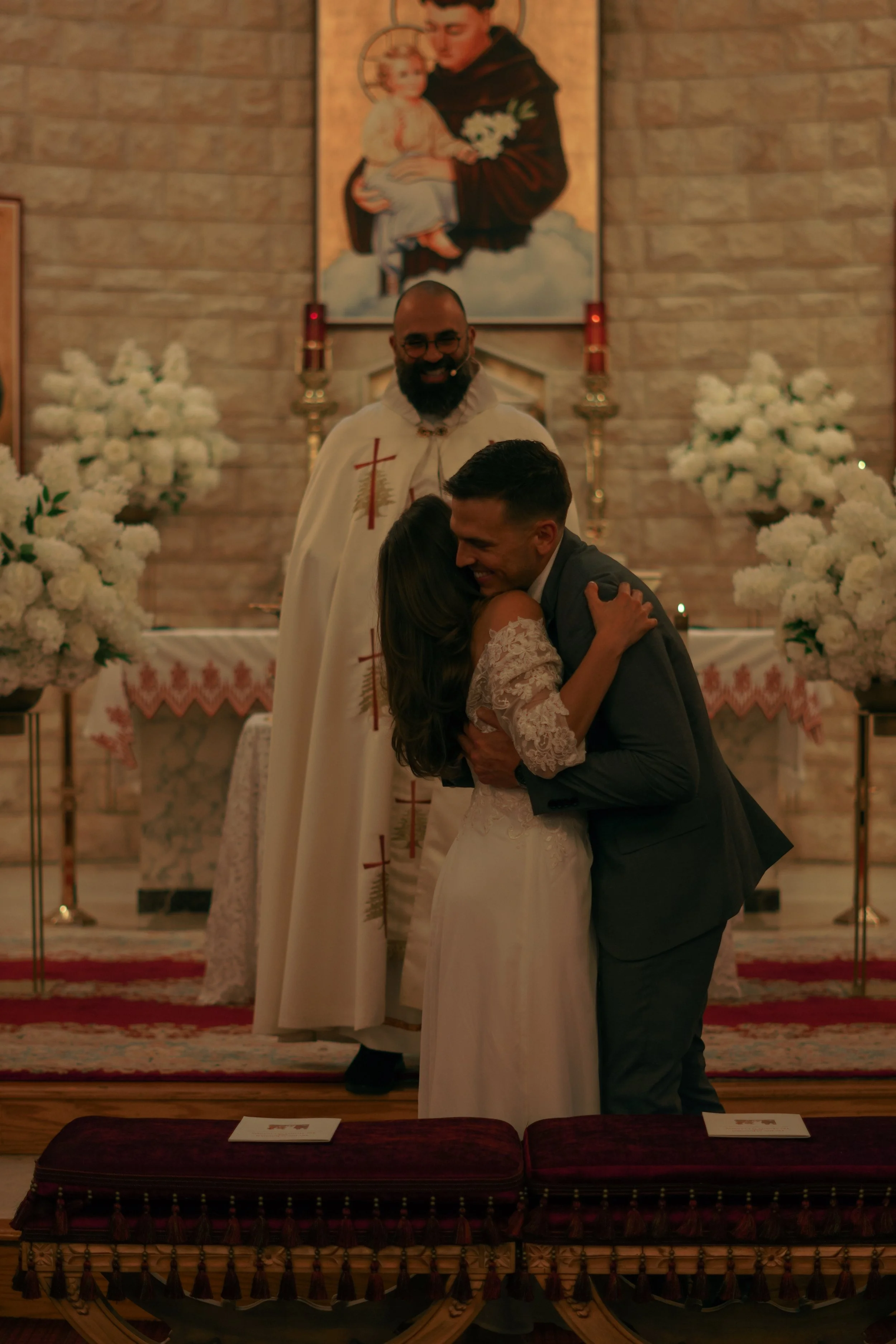 A wedding ceremony inside a church with a bride and groom embracing, a priest smiling behind them, and an altar decorated with white flowers and religious iconography.
