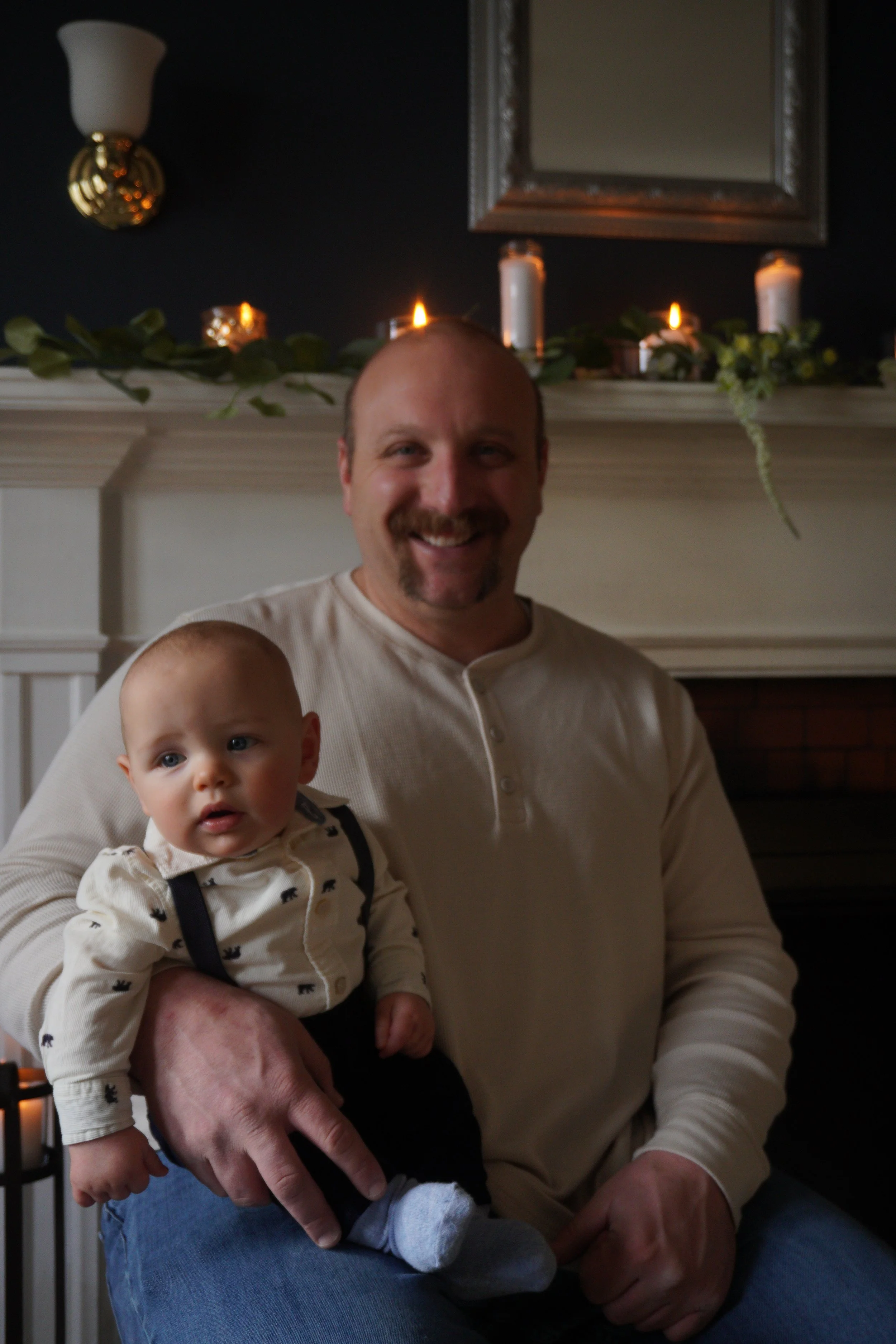 A man with a mustache and goatee holding a baby. The man is smiling and wearing a light-colored shirt. The baby has light-colored hair, blue eyes, and is dressed in a cream-colored shirt with a black animal pattern and suspenders. The background incl