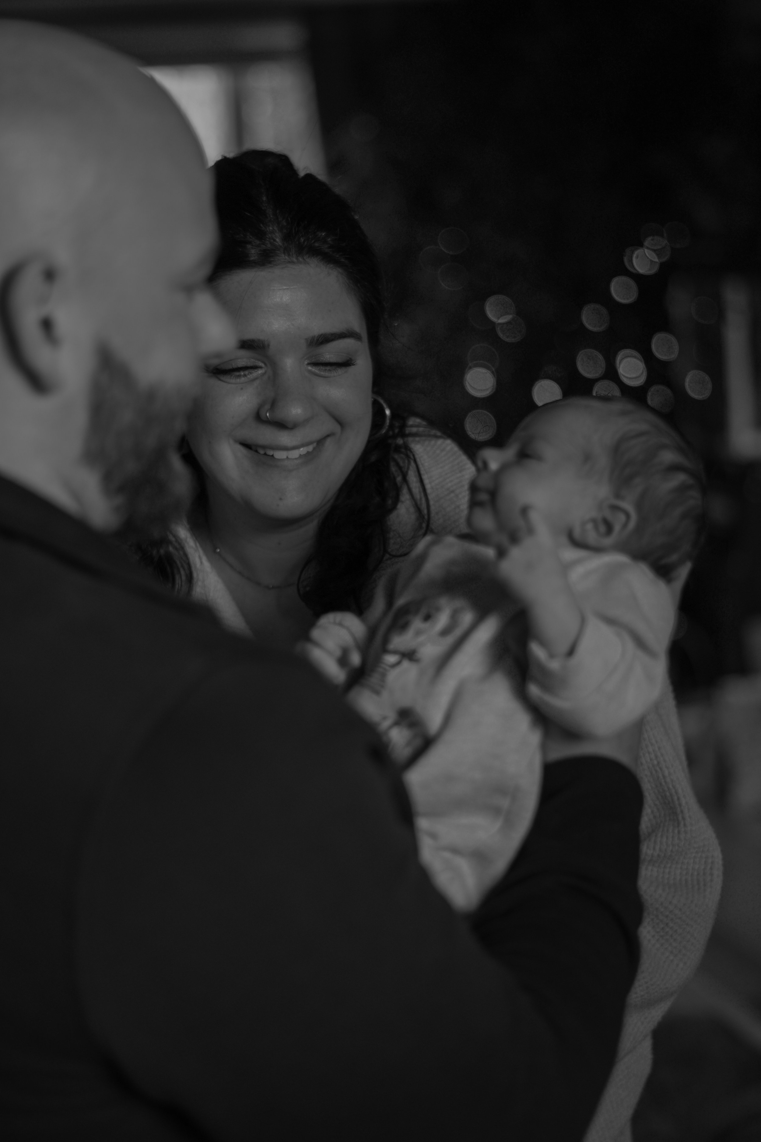 A woman with closed eyes smiling, holding a baby with light-colored hair, in a dark environment with blurred lights in the background, black and white photo.