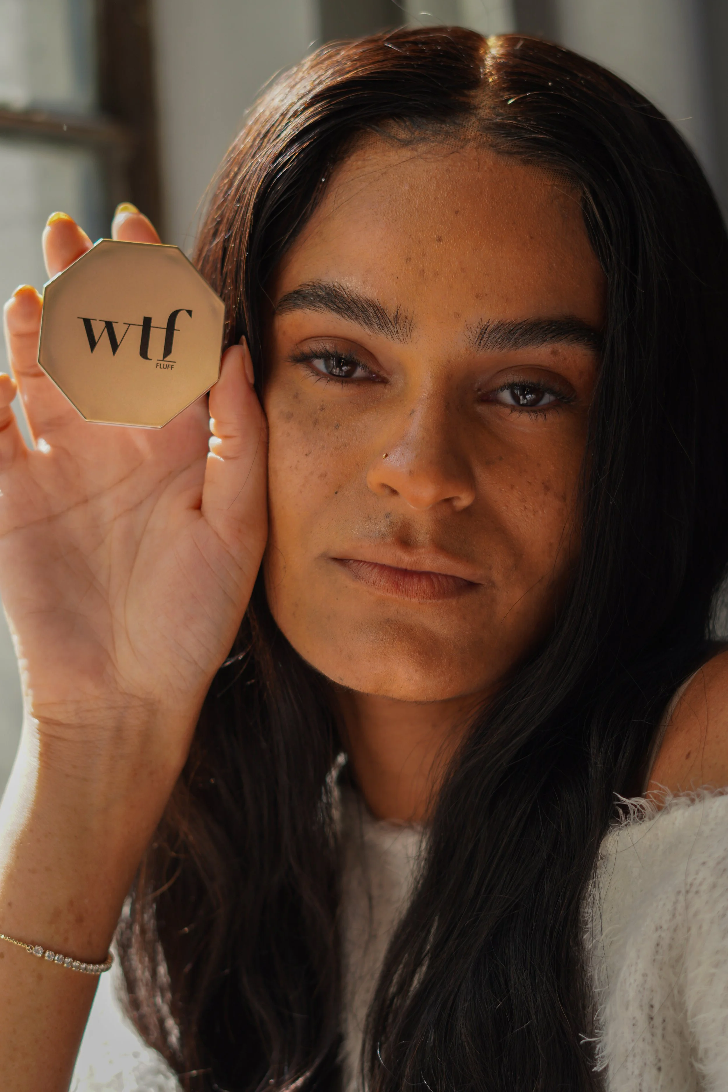 A woman with long dark hair holding a compact mirror labeled 'wlf FLUFF' up to her face. She is wearing a white fluffy garment, a bracelet on her wrist, and has a small nose piercing. The background appears to be indoors with natural light.