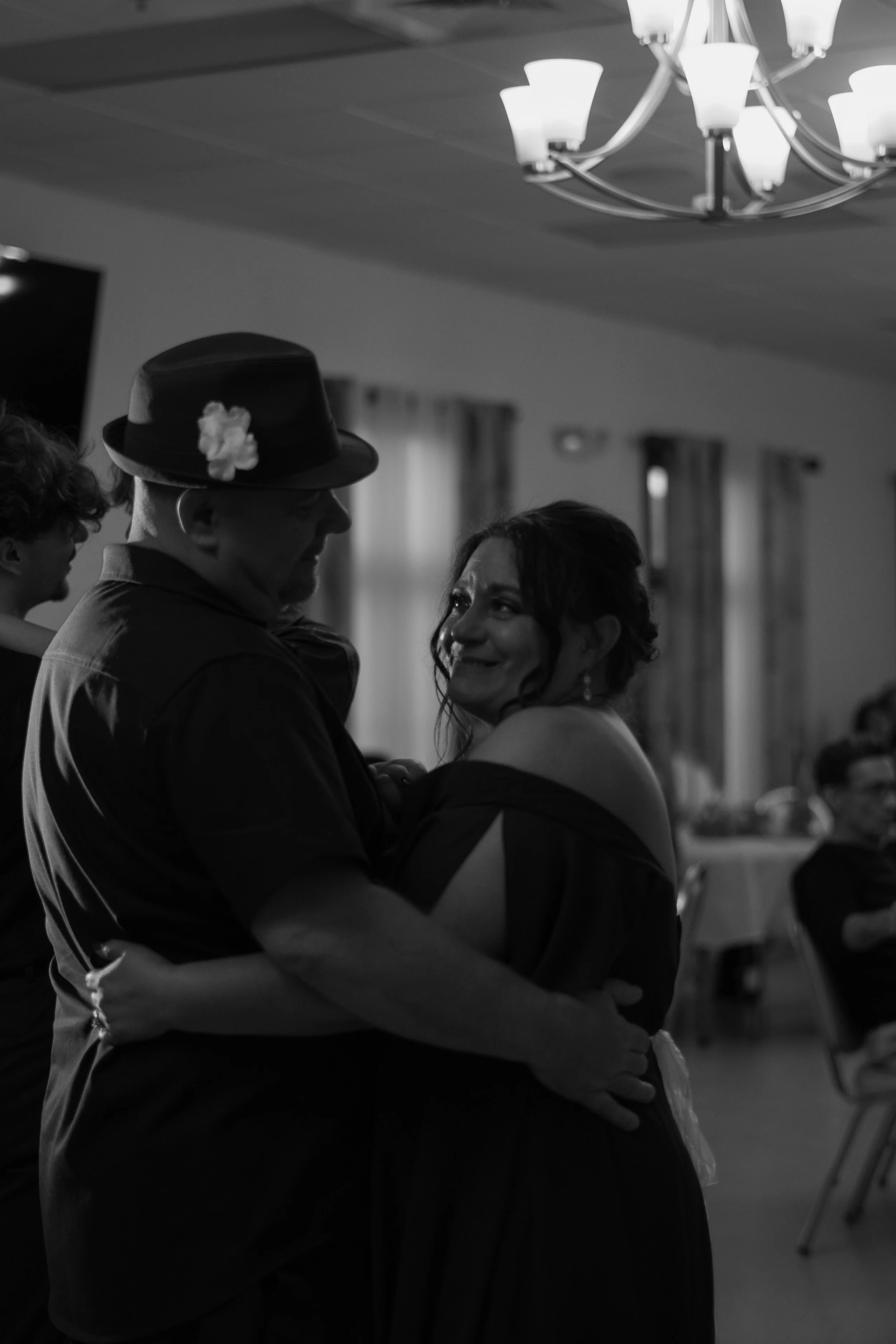 A couple dancing closely at a social event, with the man wearing a fedora hat decorated with a flower and the woman smiling at him, in a room with curtains and a chandelier.