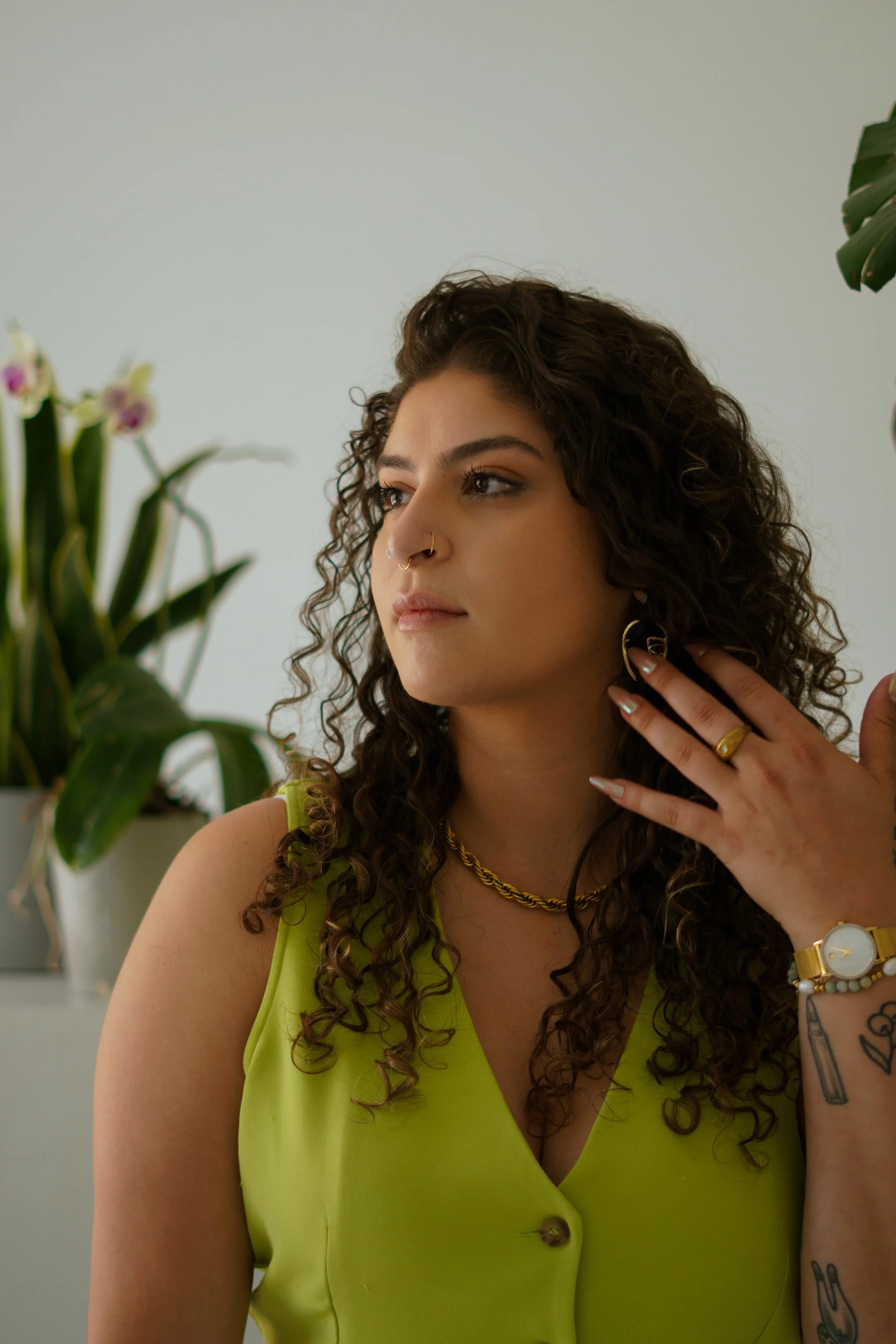 A woman with curly dark hair wearing a neon green top, gold jewelry, and multiple rings, touching her neck, with a potted plant and orchids in the background.