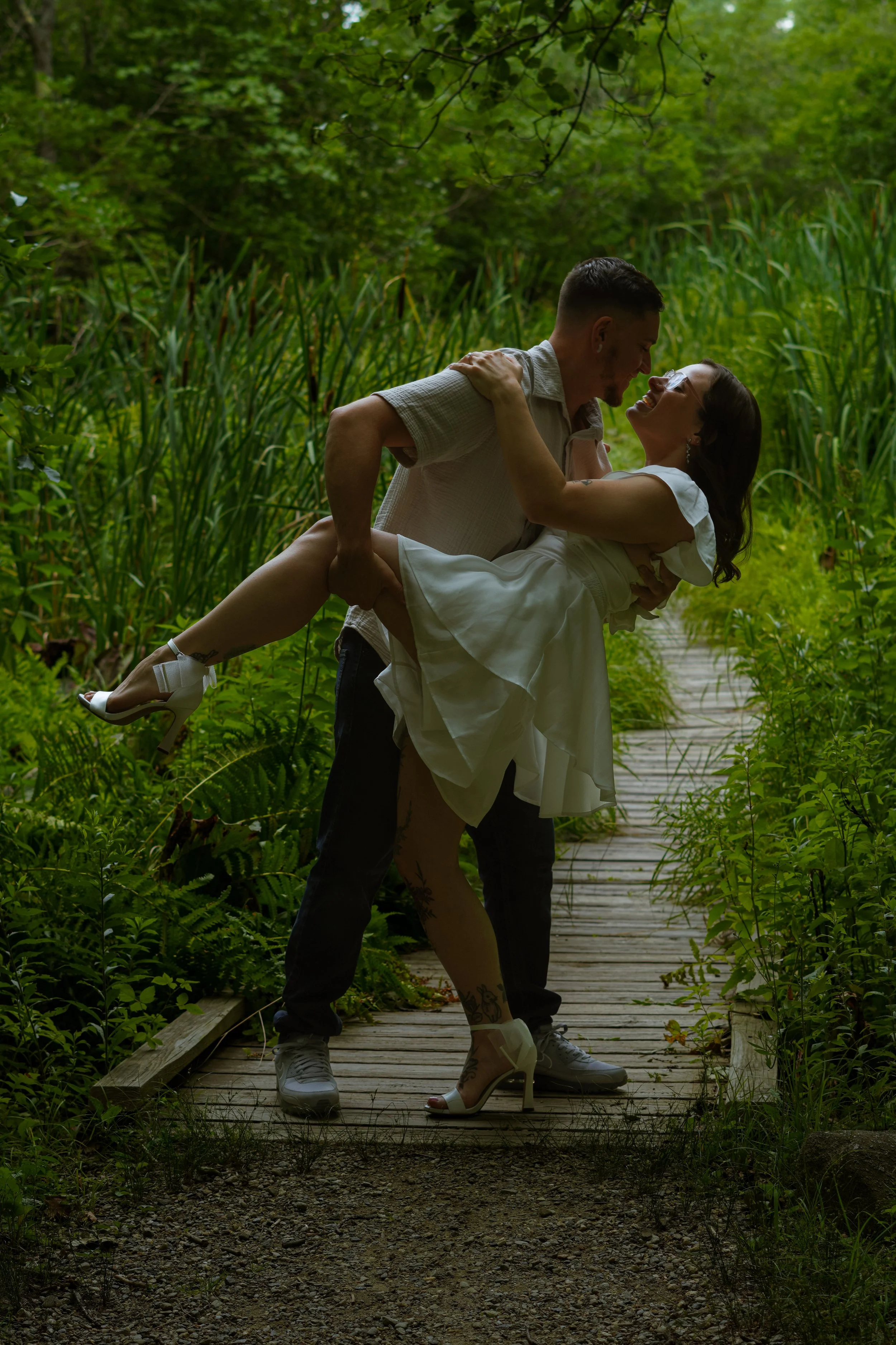 A man holding a woman in a white dress and high heels on a wooden pathway surrounded by lush green foliage.