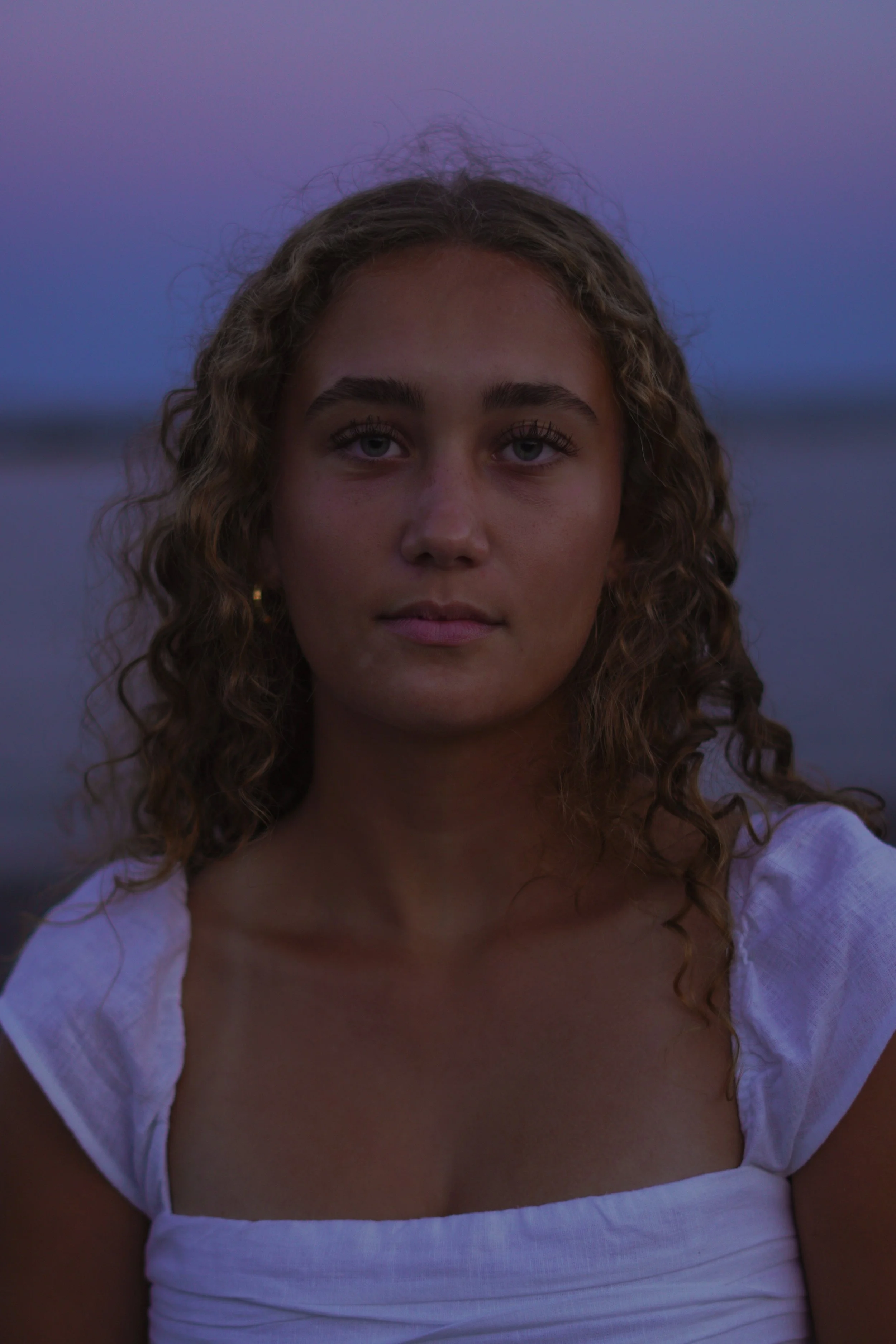 A young woman with curly brown hair and blue eyes wearing a white sundress hanging off her shoulders, standing outdoors at dusk or dawn with a blurred water and sky background.