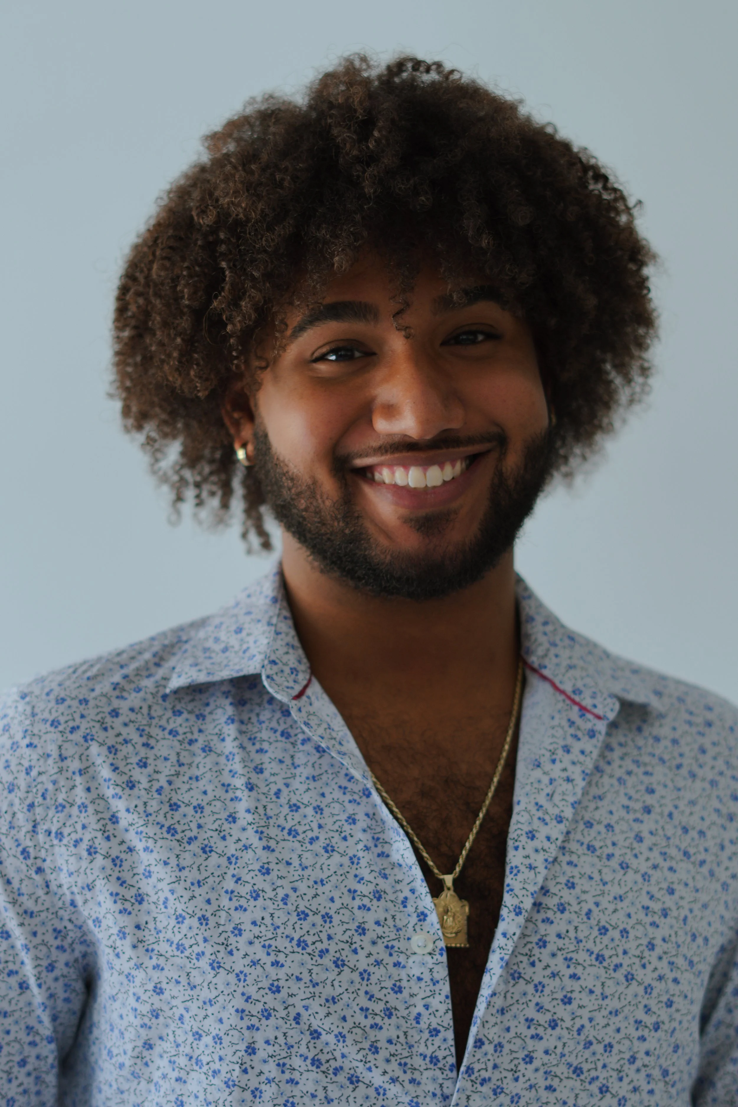 A smiling man with curly hair, a beard, earrings, and wearing a patterned button-up shirt and gold jewelry, standing against a plain light blue background.