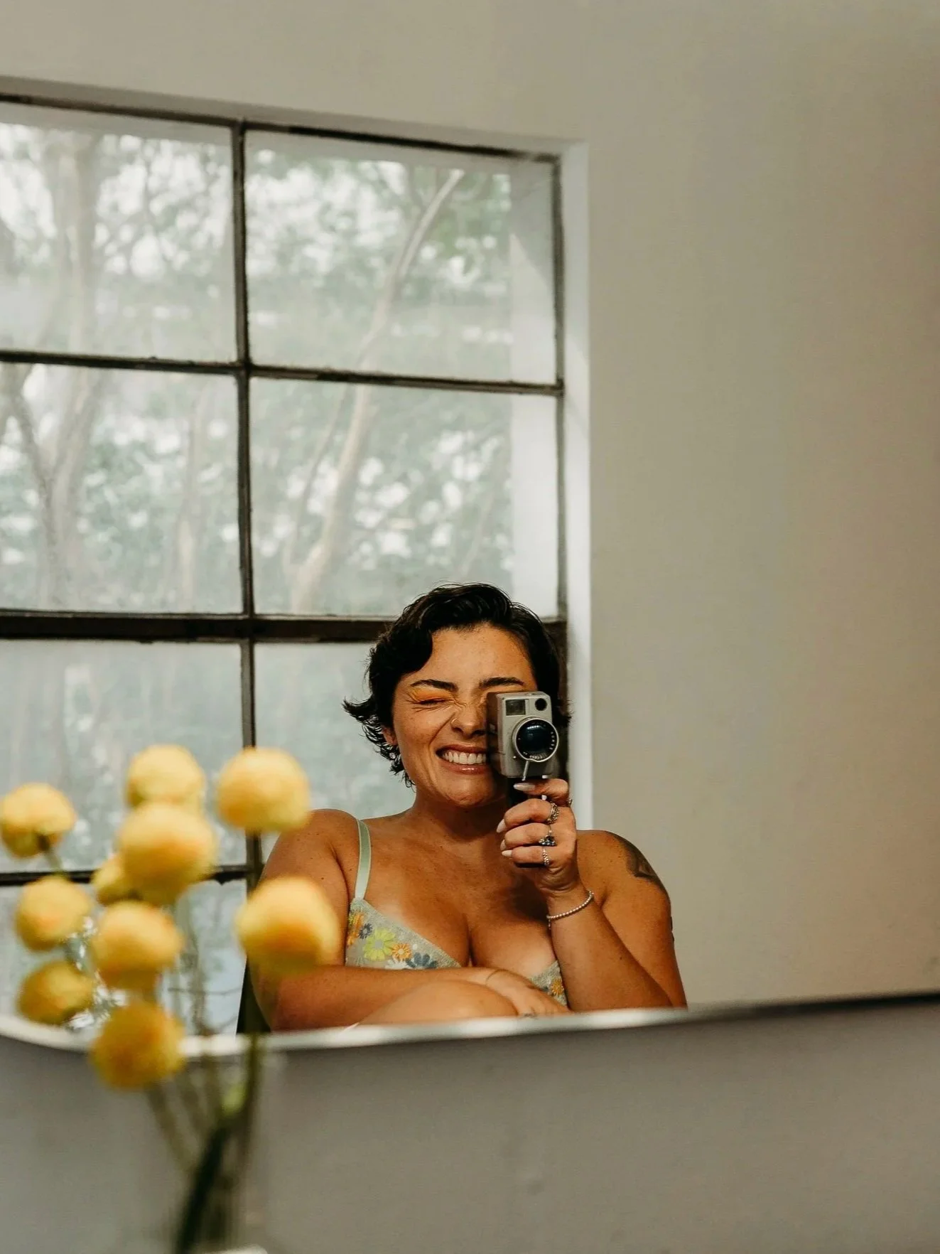 A woman is taking a selfie in a mirror, smiling and winking, with a window showing trees in the background. She is holding a camera and has short dark hair, wearing a sleeveless top.