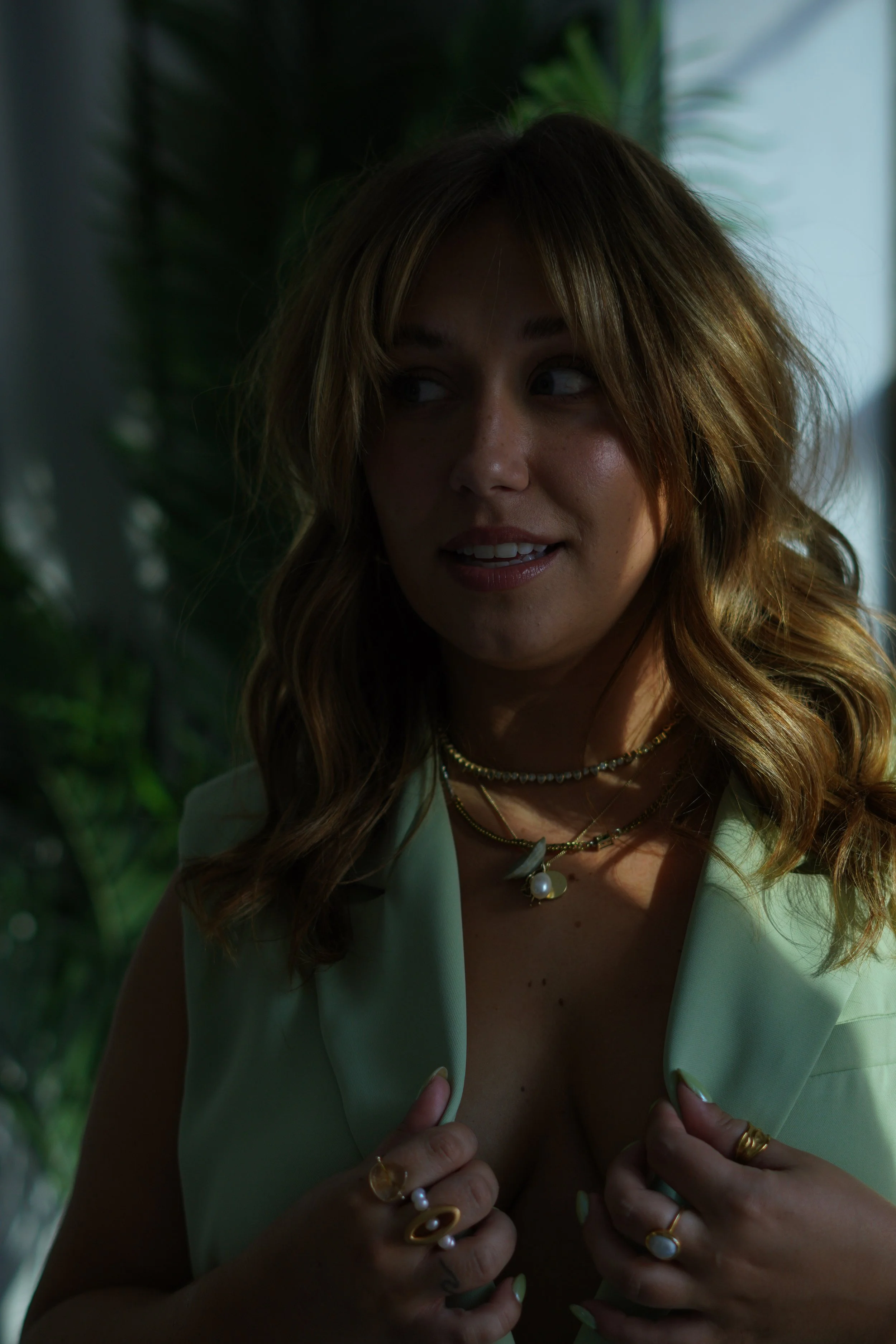 A woman with wavy, light brown hair looking to the side, smiling slightly, wearing a light-colored sleeveless top and multiple gold jewelry pieces including rings, necklaces, and earrings, standing in front of a leafy green background with sunlight f