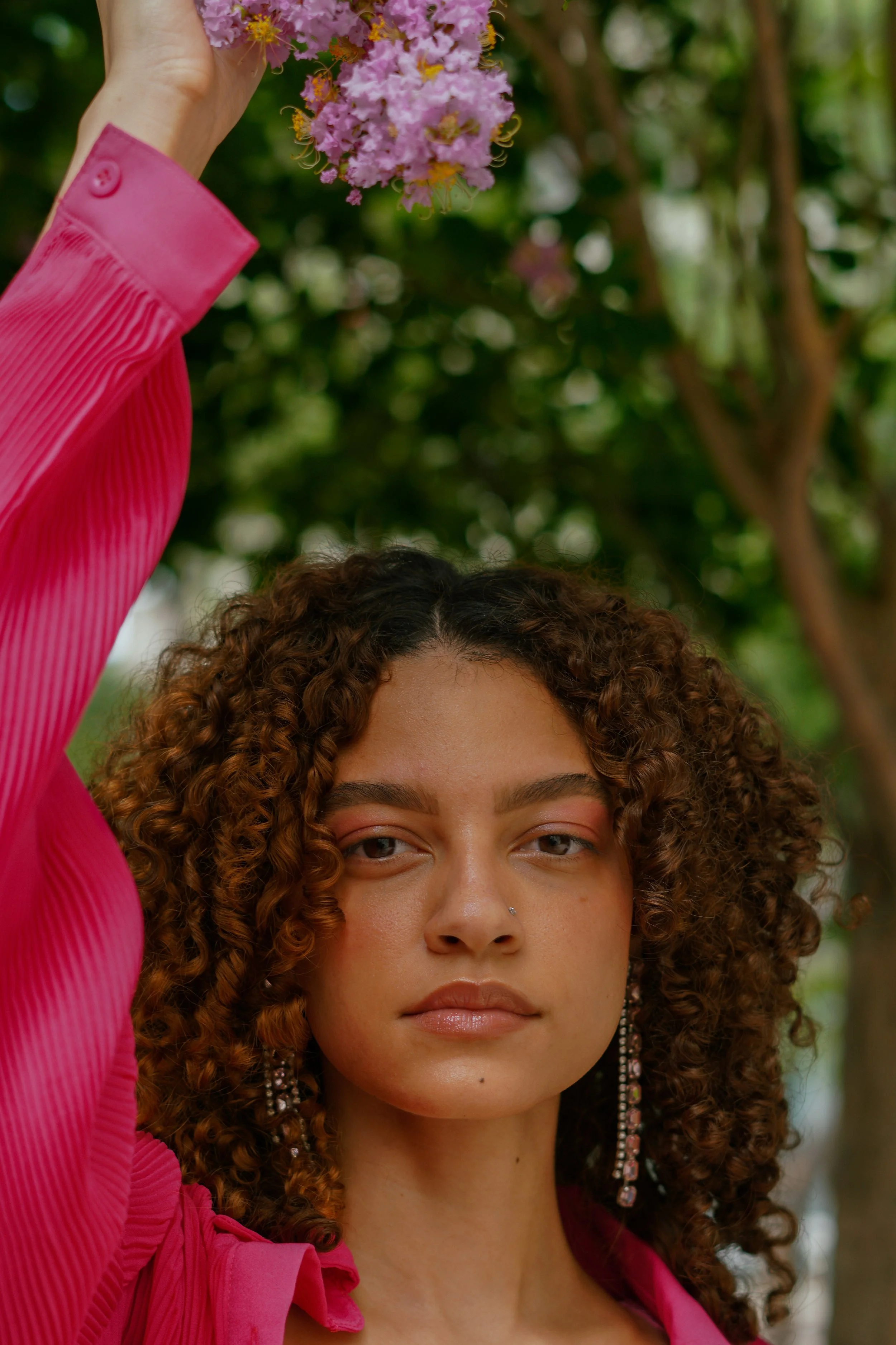 A woman with curly hair and a nose ring, wearing a pink top and long earrings, holding a pink flower in front of her forehead outdoors with trees in the background.