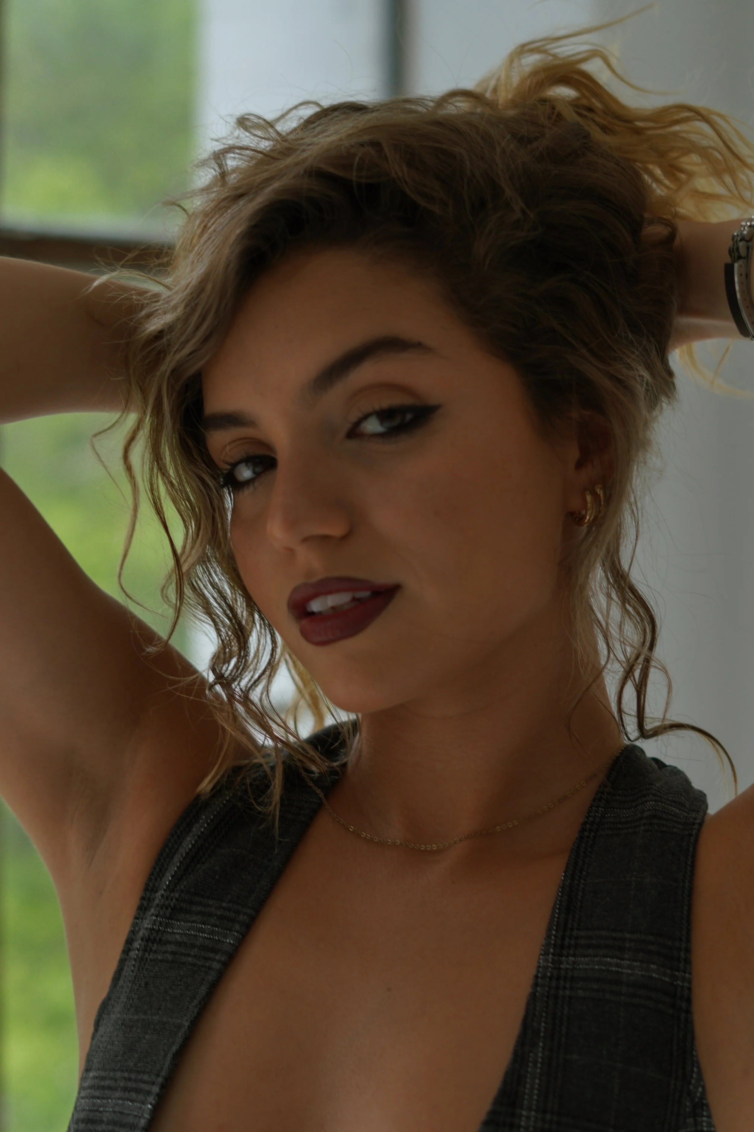 A young woman with curly hair and winged eyeliner posing indoors, wearing a sleeveless dark plaid dress and a gold necklace.