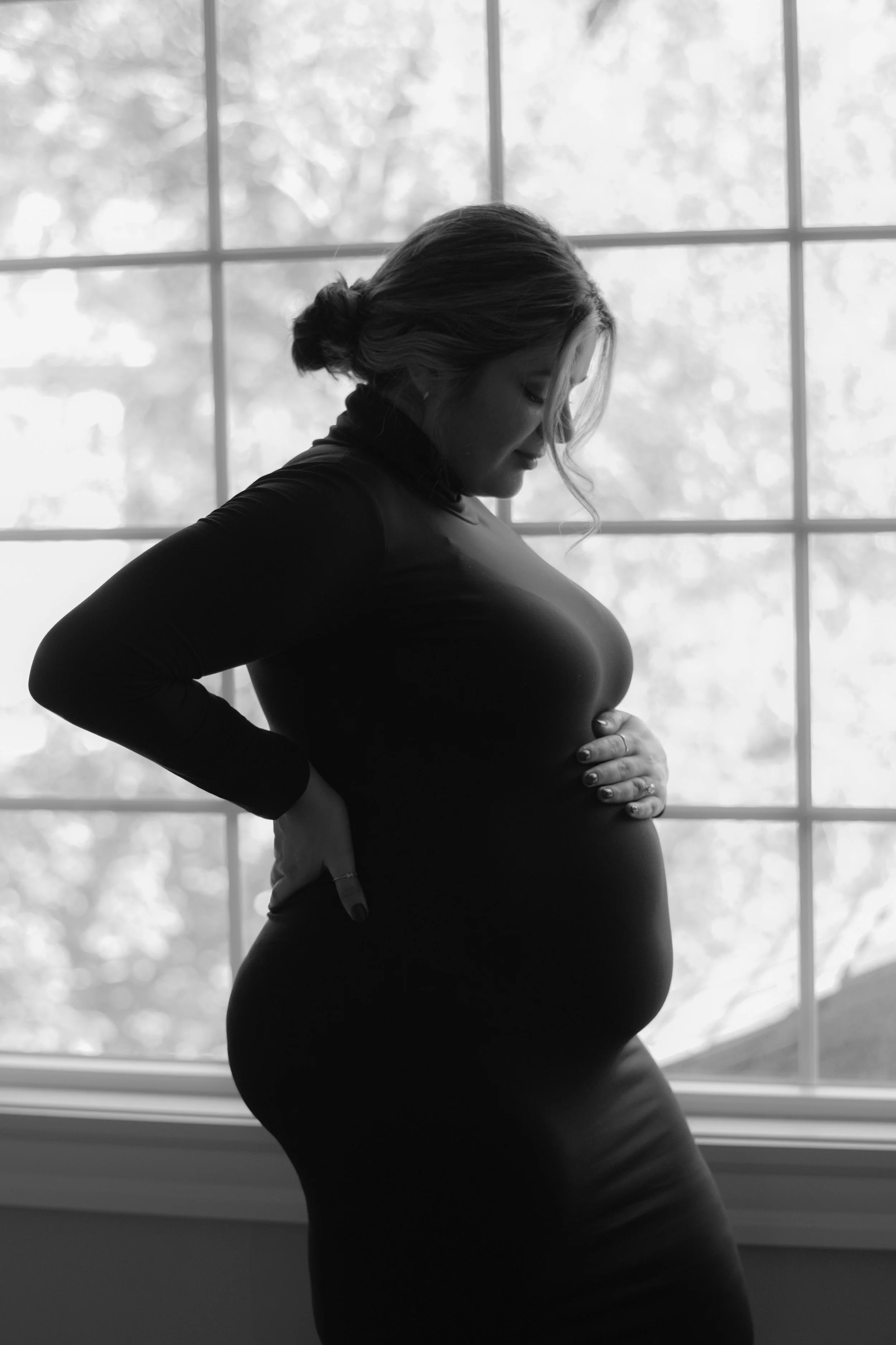 Black and white photo of a pregnant woman standing indoors near a large window, gently holding her baby bump with both hands, looking down with a serene expression.