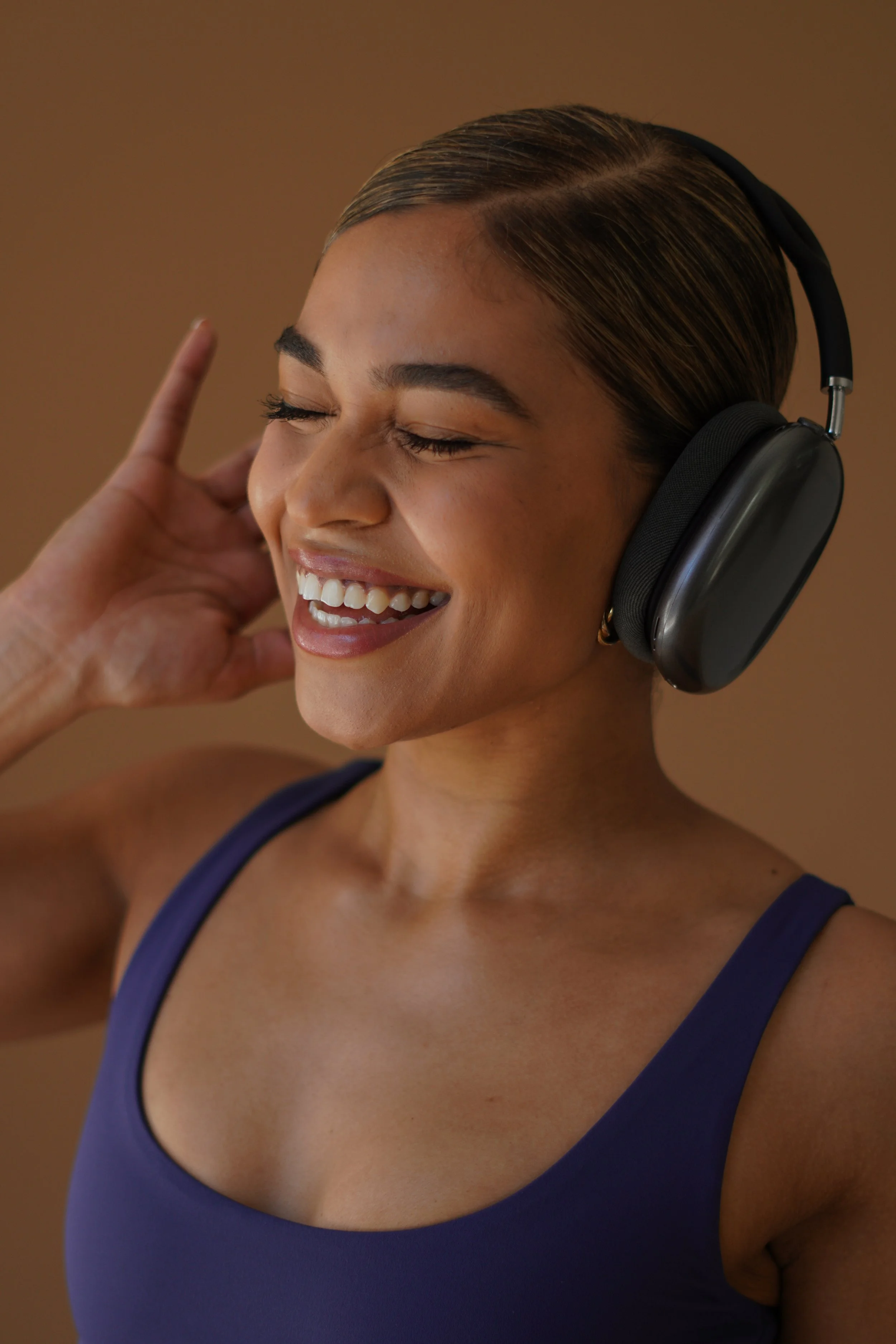 A woman with brown hair and closed eyes wearing black headphones and a blue tank top, smiling with her hand near her ear.