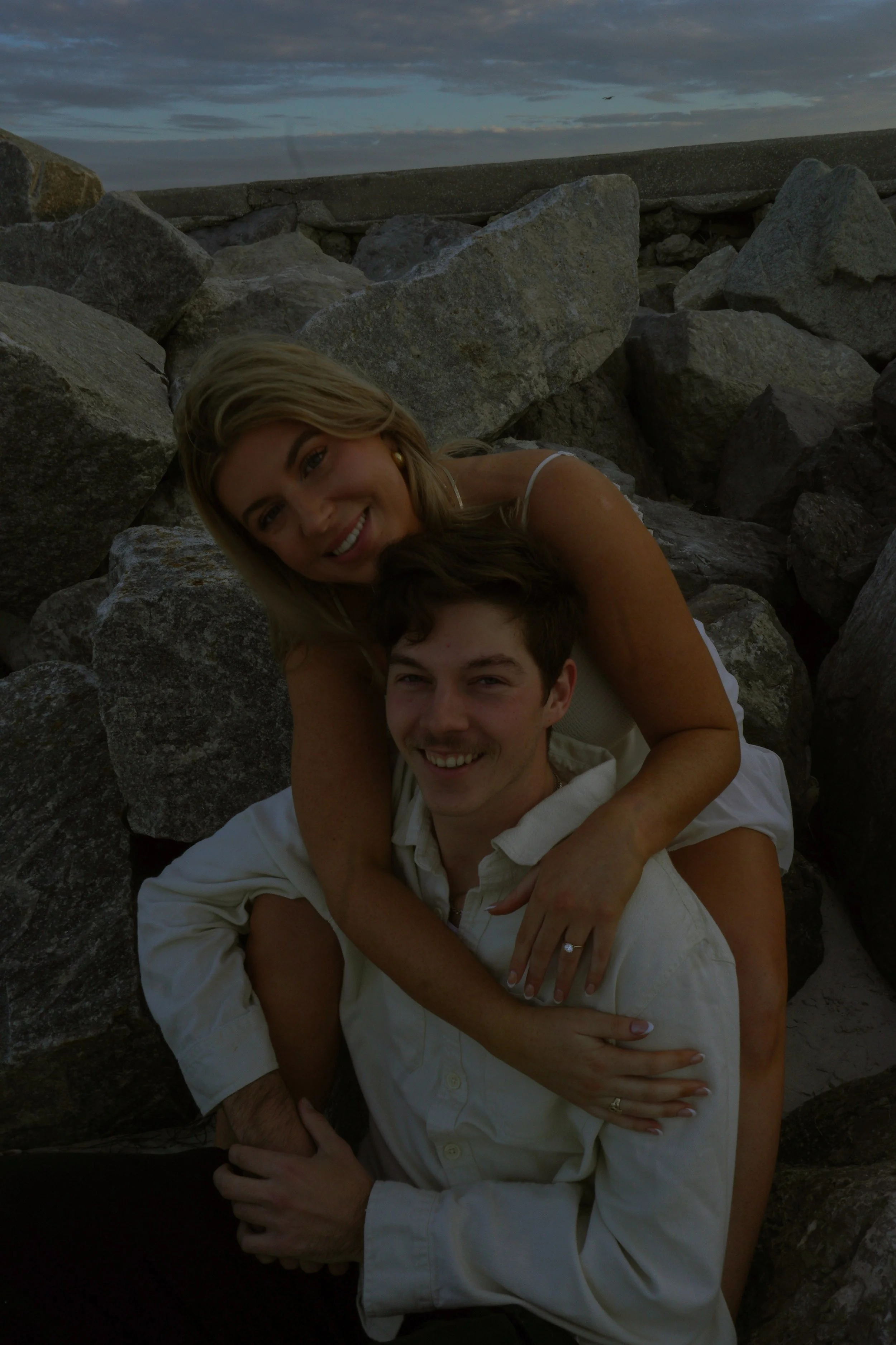 Two smiling people, a woman with blonde hair and a man with dark hair, embracing each other among large rocks outdoors during sunset or dusk.