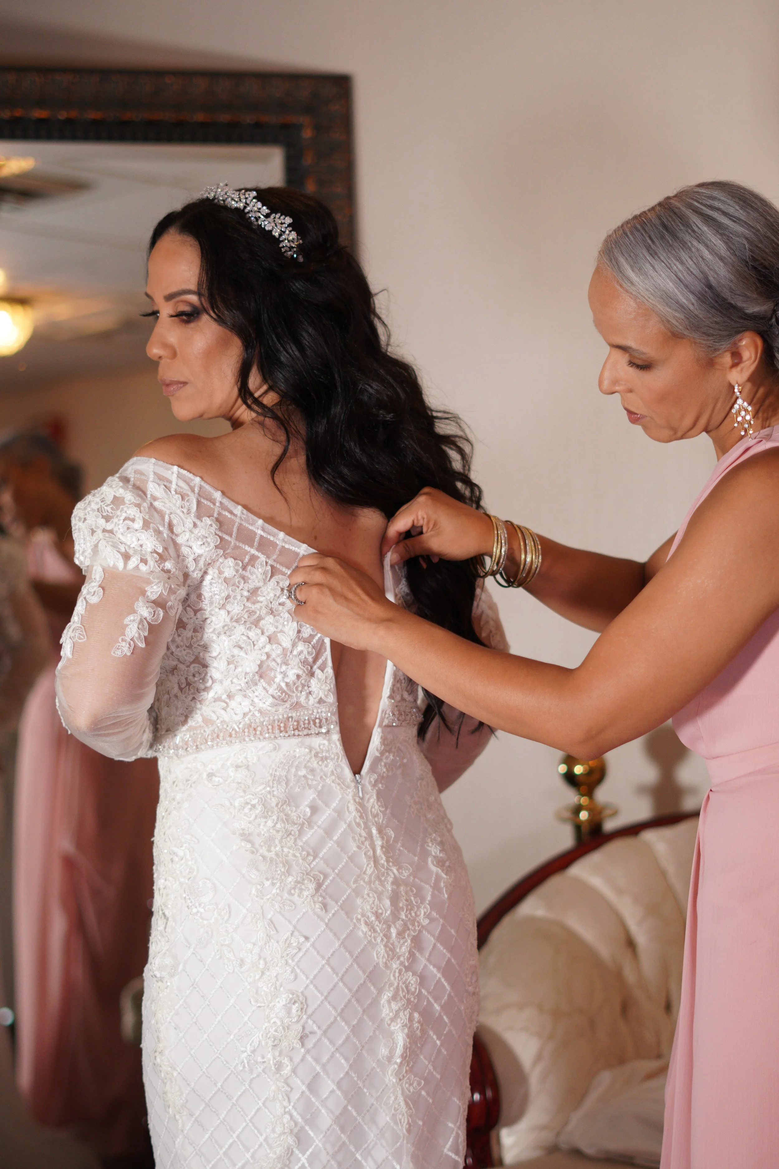 A bride in an elegant white lace wedding dress with floral embroidery gets ready with assistance from a woman in a pink dress, likely her mother, in a warmly lit room decorated with a mirror and a beige armchair.