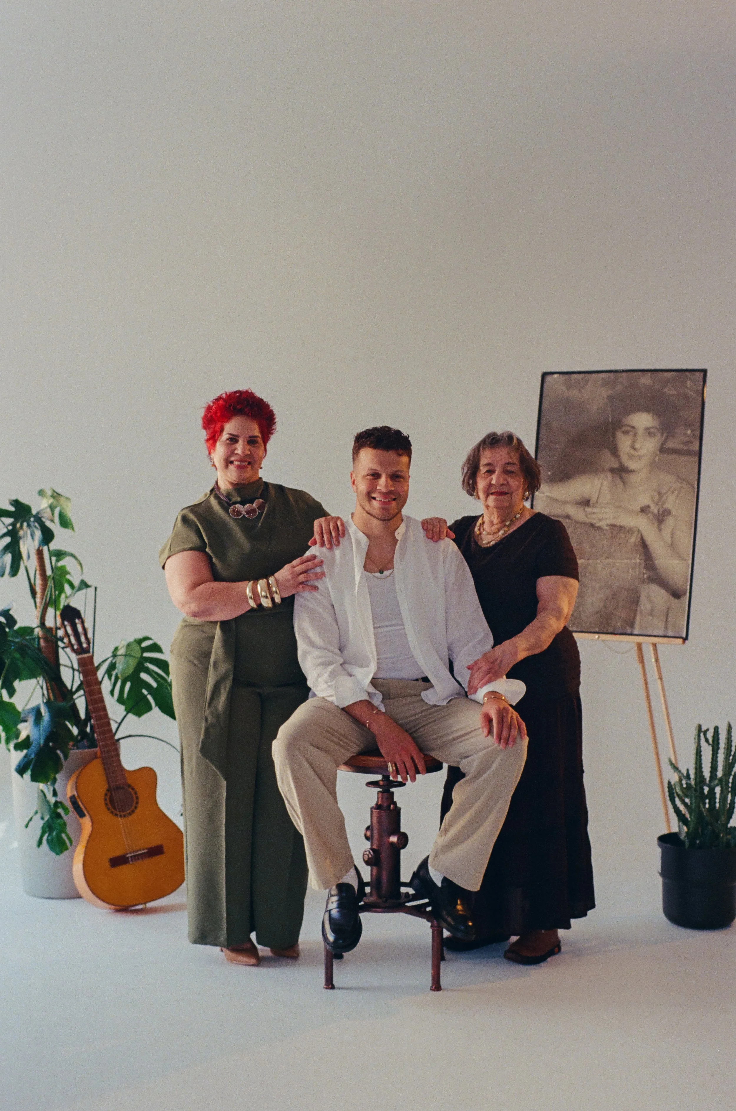 Three people posing together, one sitting on a stool, two standing beside. There is a guitar and potted plants nearby, and a photo of a woman on an easel in the background.