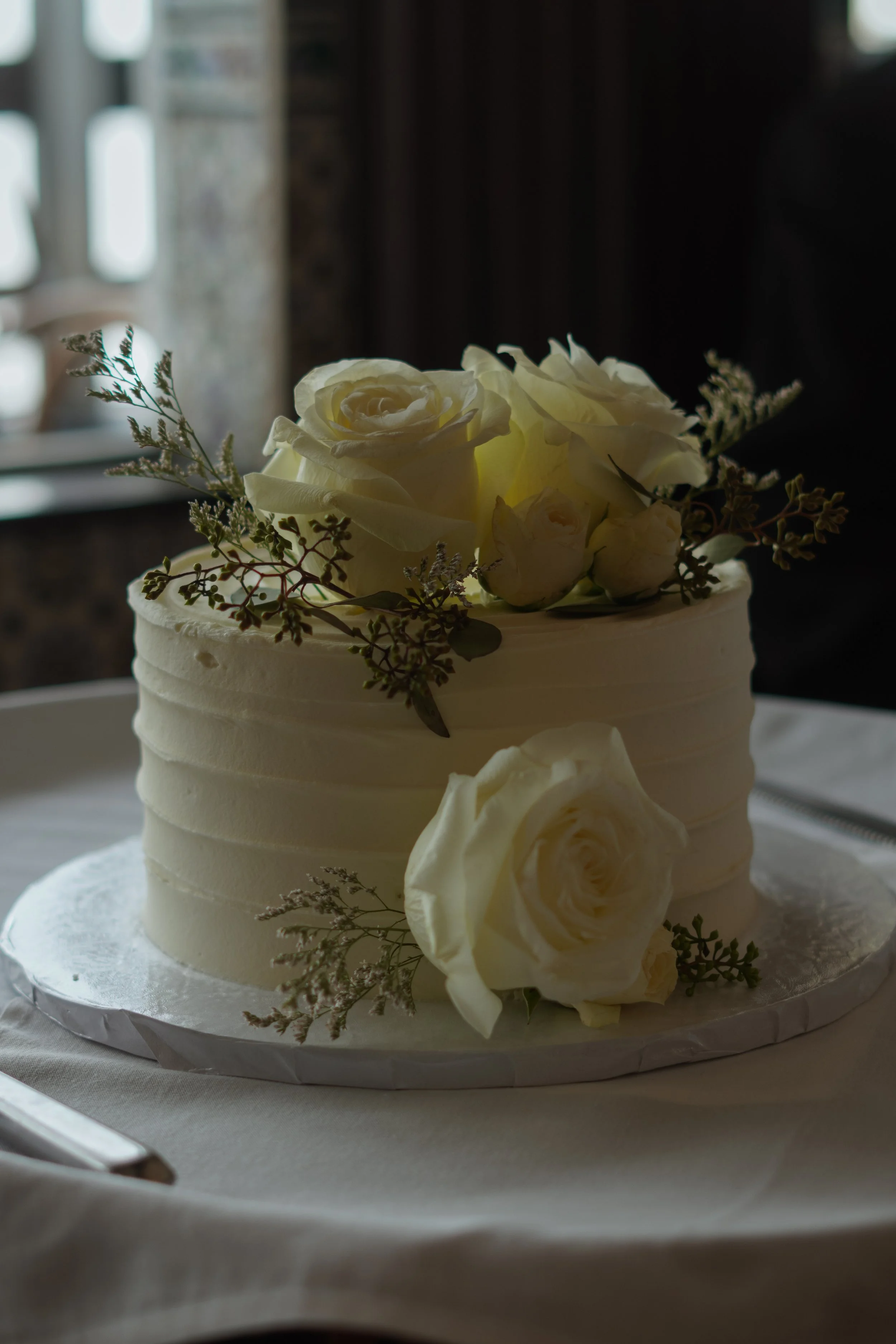 A white frosted wedding cake decorated with white roses and greenery, placed on a white cake board on a table.