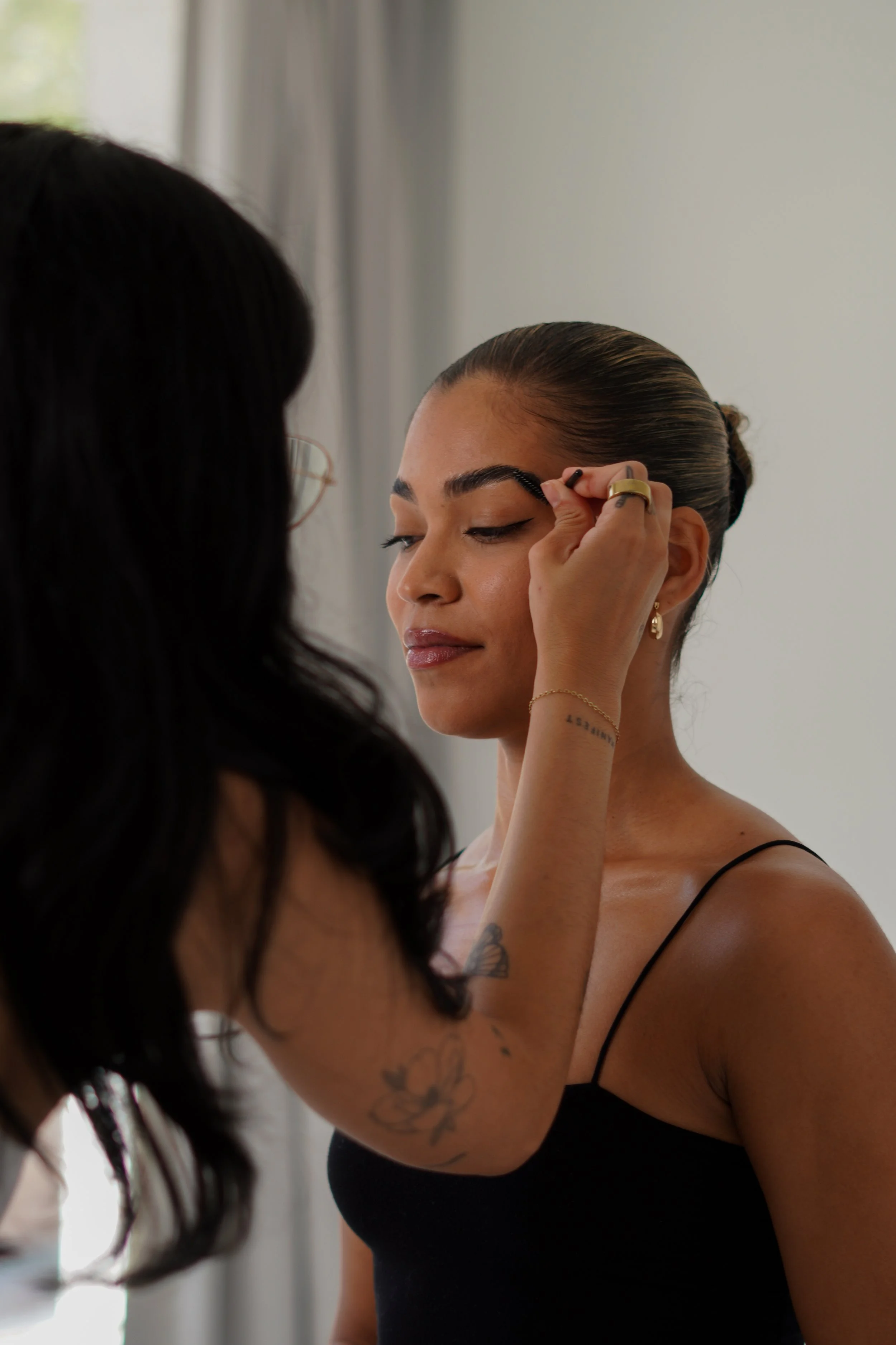 A woman with her hair tied back is getting her makeup done by another woman with long dark hair and glasses, inside a room with white walls and a window.