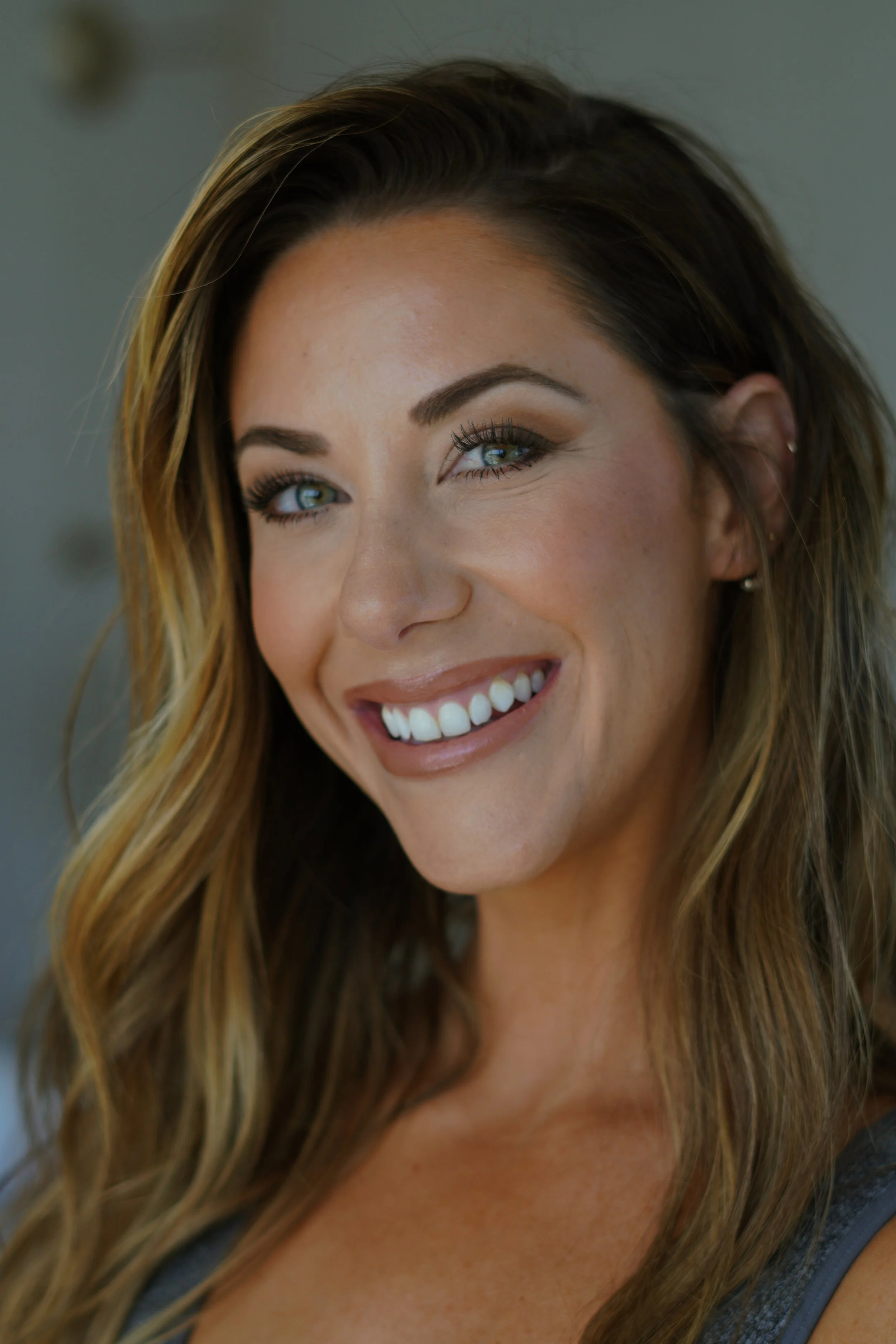 Close-up of a woman with long, wavy blonde hair, blue eyes, and makeup, smiling, with a blurred background.