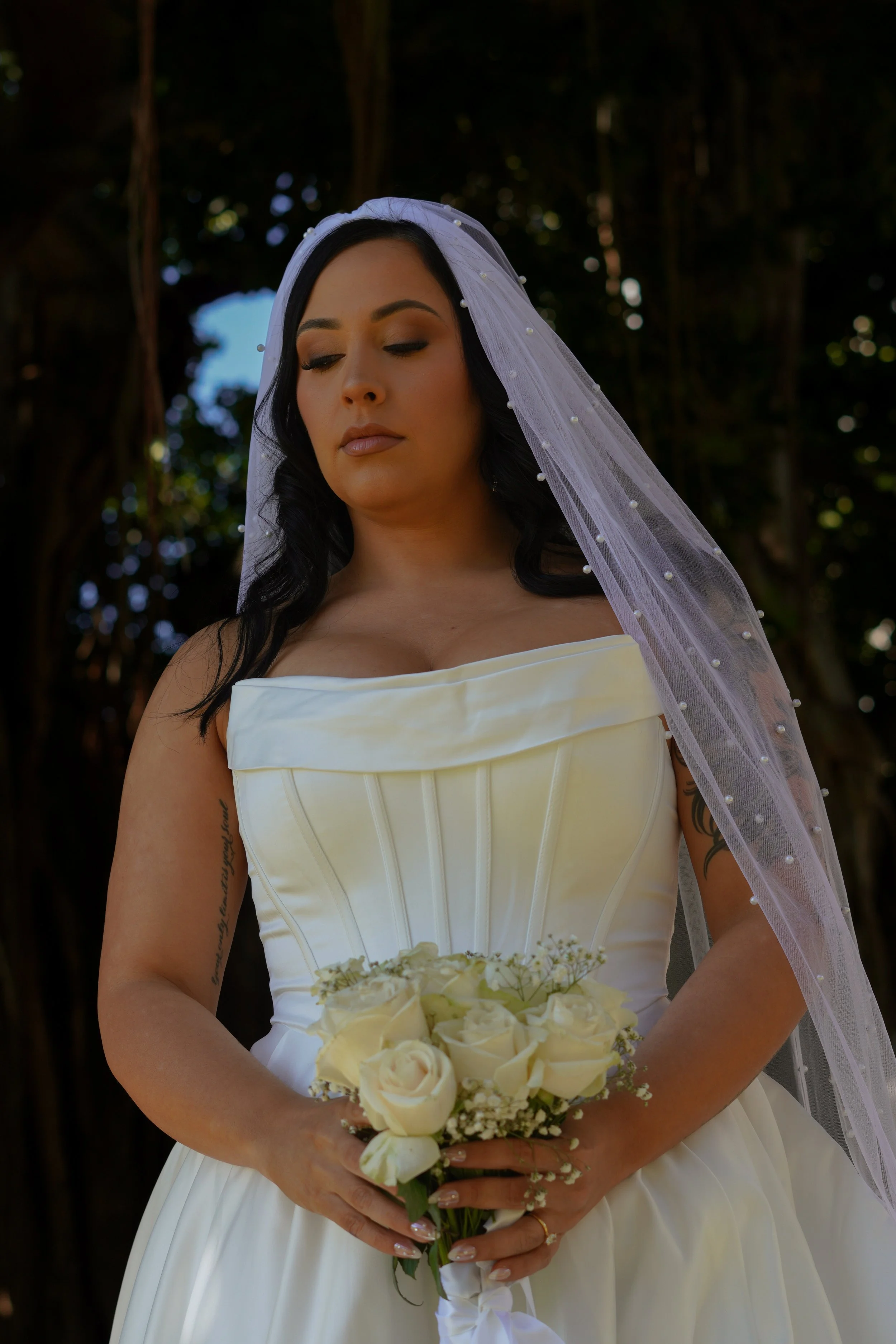 A woman in a wedding dress holding a bouquet of white roses and flowers, with a veil over her head, standing outdoors in front of trees.