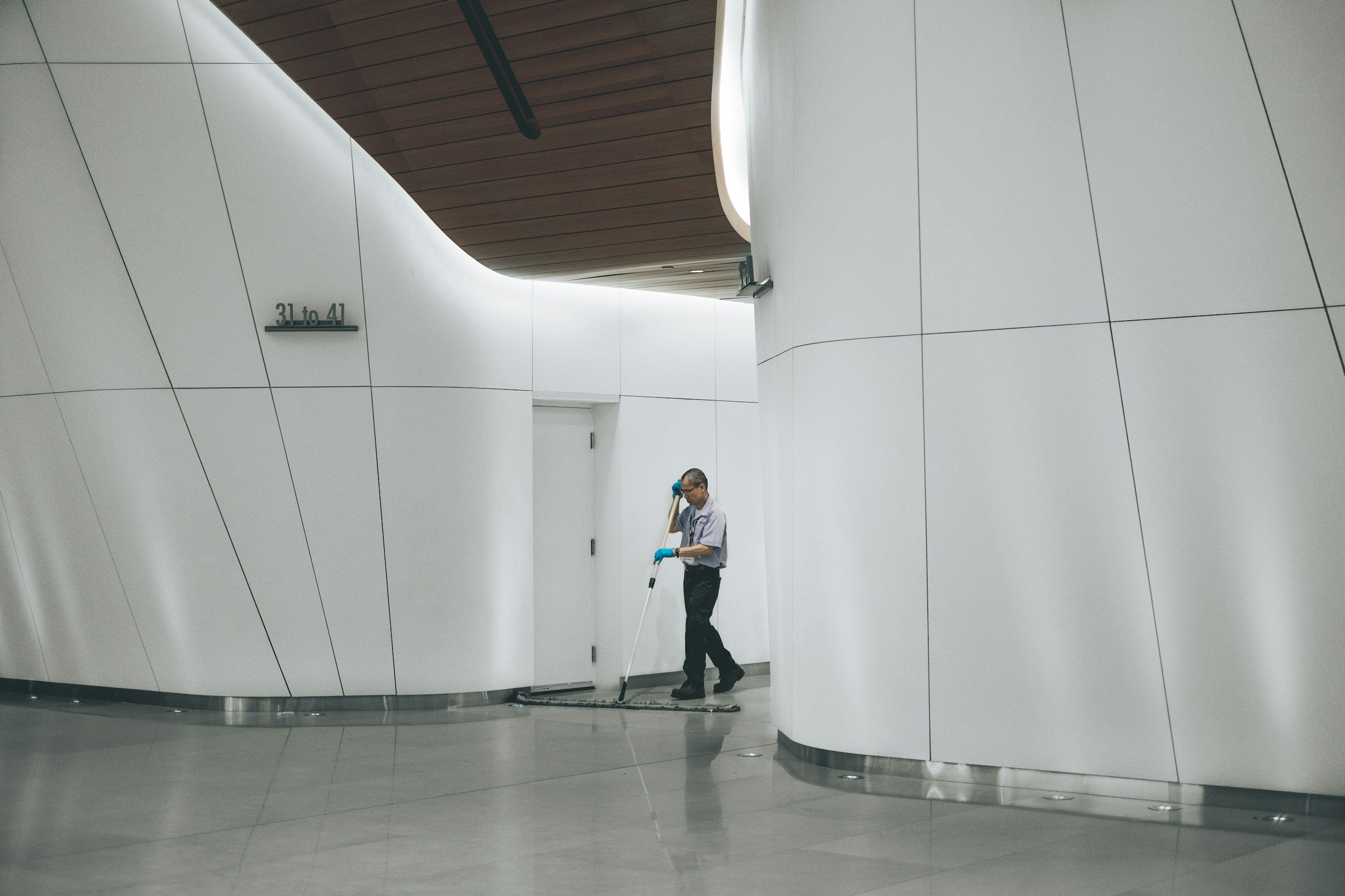 A person wearing glasses and blue gloves cleaning the floor of a modern, minimalist corridor with white curved walls and a wooden ceiling.