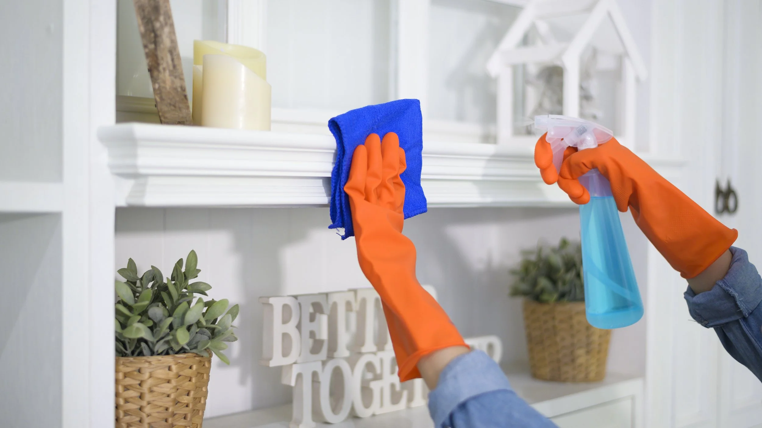 Hands wearing orange gloves cleaning a white shelf with a blue spray bottle and a blue cloth, with candles and decorative items in the background.