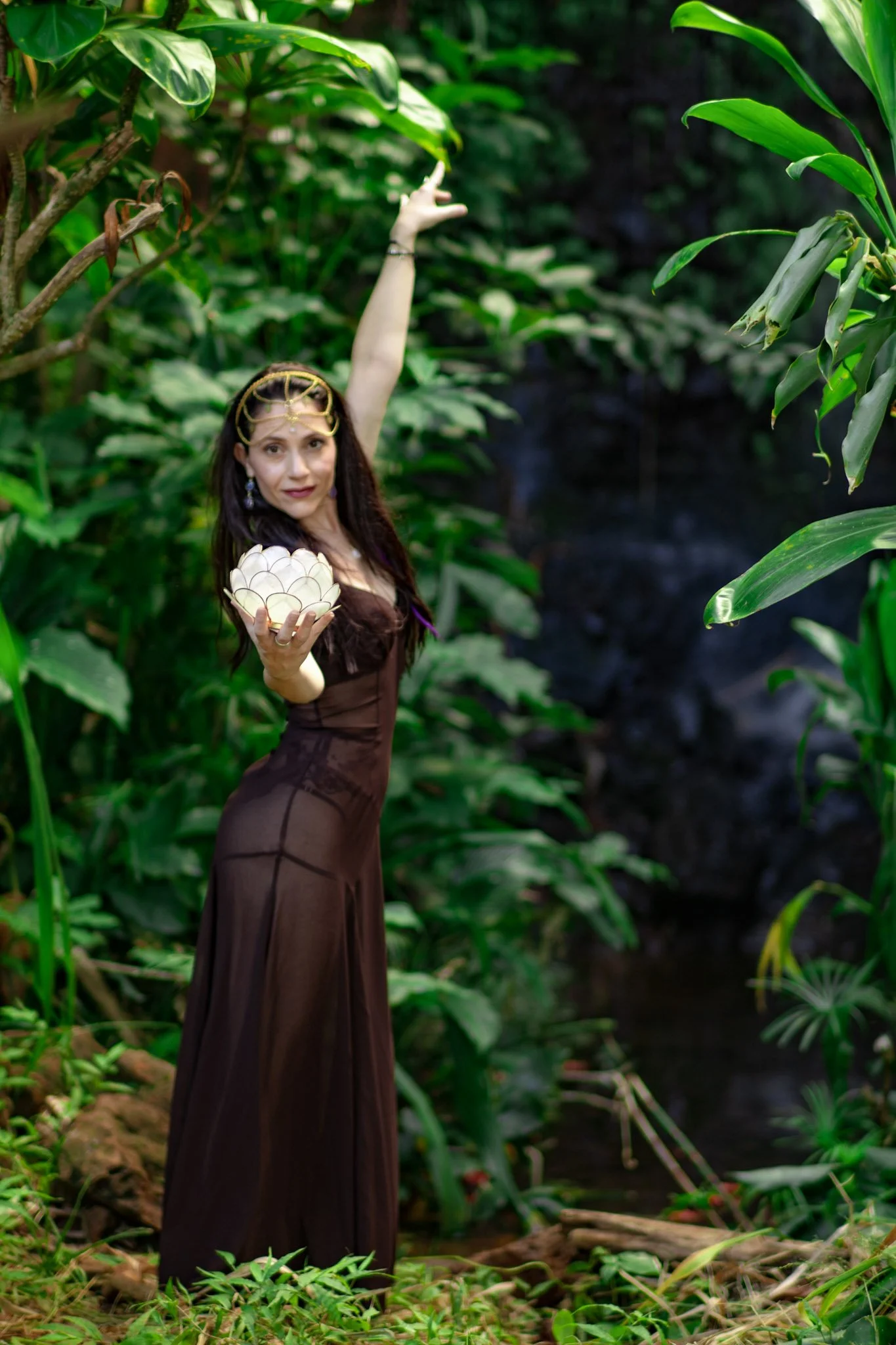 A woman dressed in a brown gown with sheer fabric standing in a lush green garden, holding a glowing lotus-shaped object and raising her arm expressing grace.