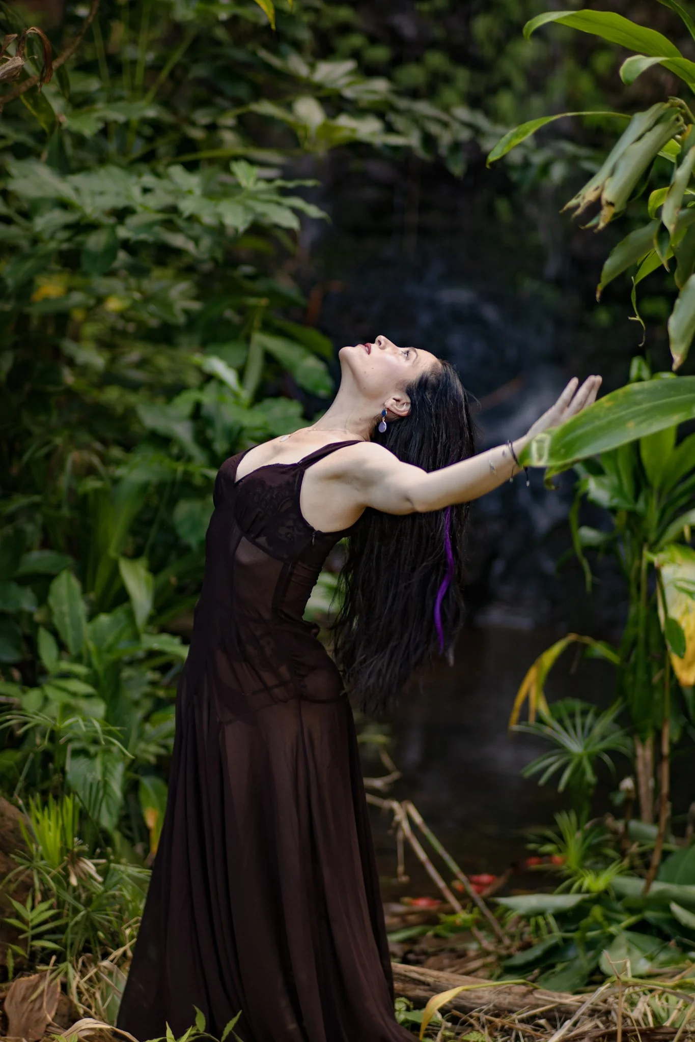 A woman with long dark hair, wearing a black dress and earrings, standing outdoors among lush green tropical plants with her arms stretched out, her head tilted back, and eyes closed in a serene pose.